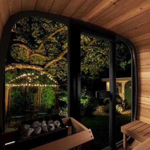 View from inside a room looking out through large glass sliding doors to an outdoor garden with a wooden bench, string lights, a tree with sprawling branches, and a house in the background.