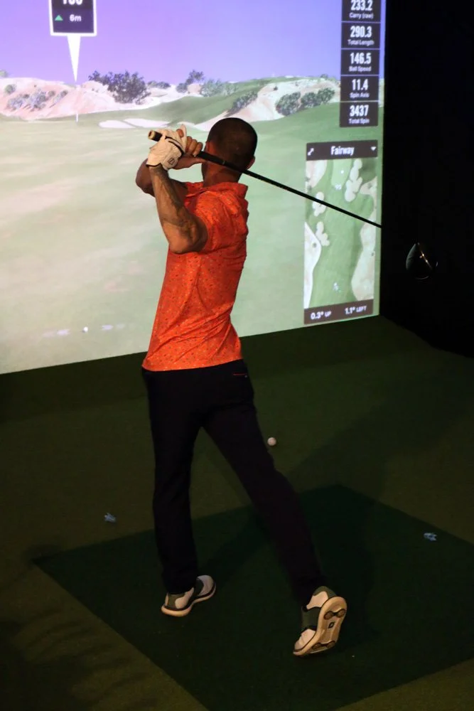 A man dressed in a red shirt and black pants practicing golf indoors on a golf simulator, holding a golf club after swinging.