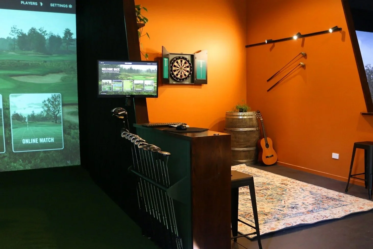 Indoor golf simulation room with a large screen displaying a golf course, a dartboard on an orange wall, a computer setup, an acoustic guitar, and golf clubs on the wall.