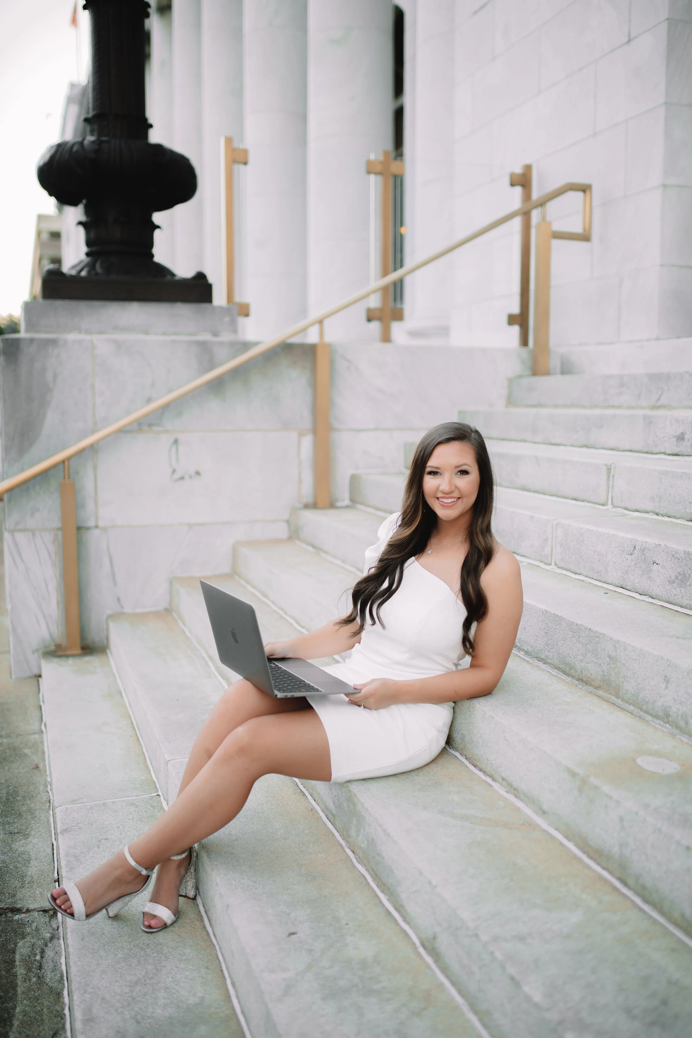 A young woman with long dark hair in a white dress sitting on stone steps outside, holding a laptop and smiling.