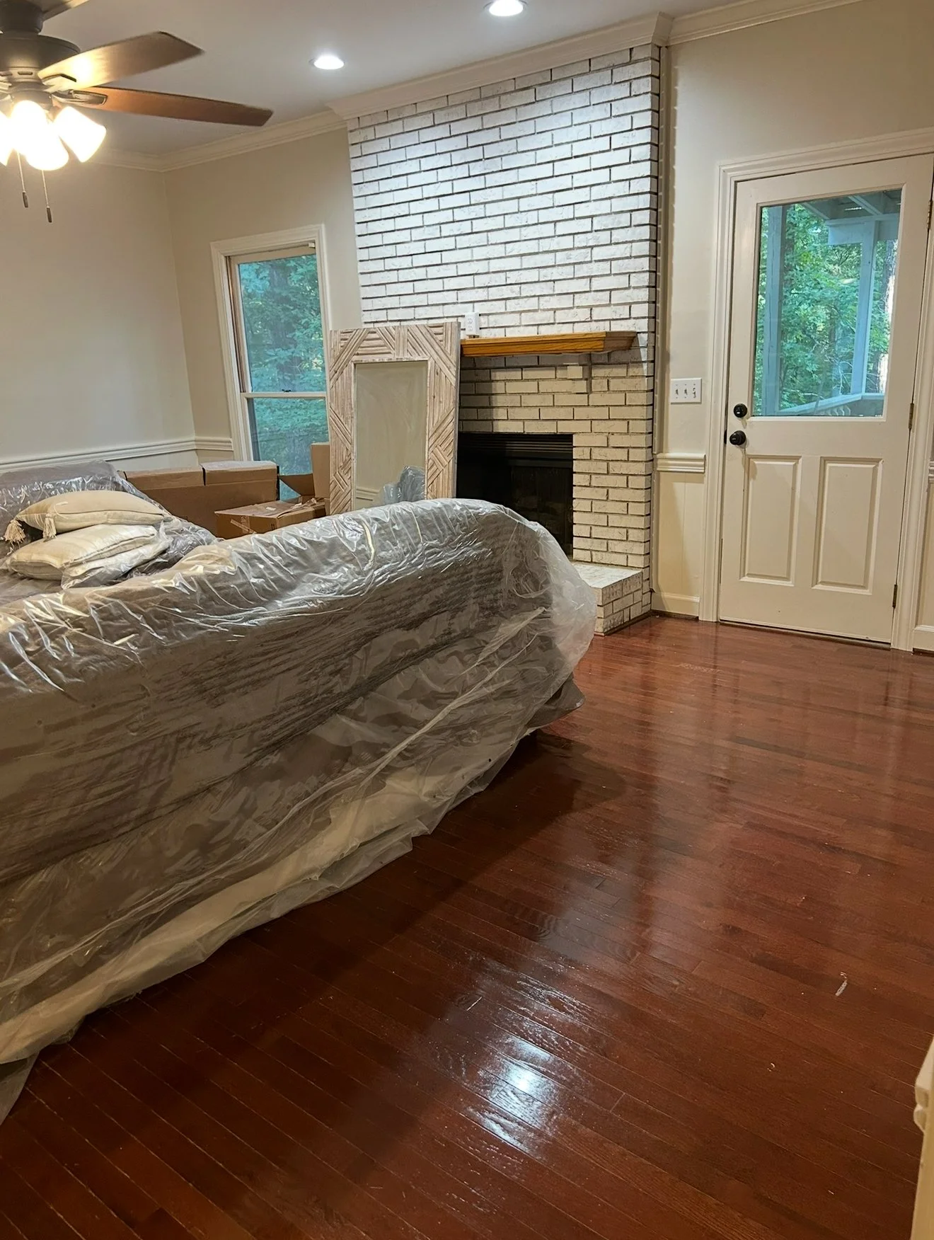 Living room with hardwood floors, a white brick fireplace, a window, a door with glass, and a beige couch covered with clear plastic.