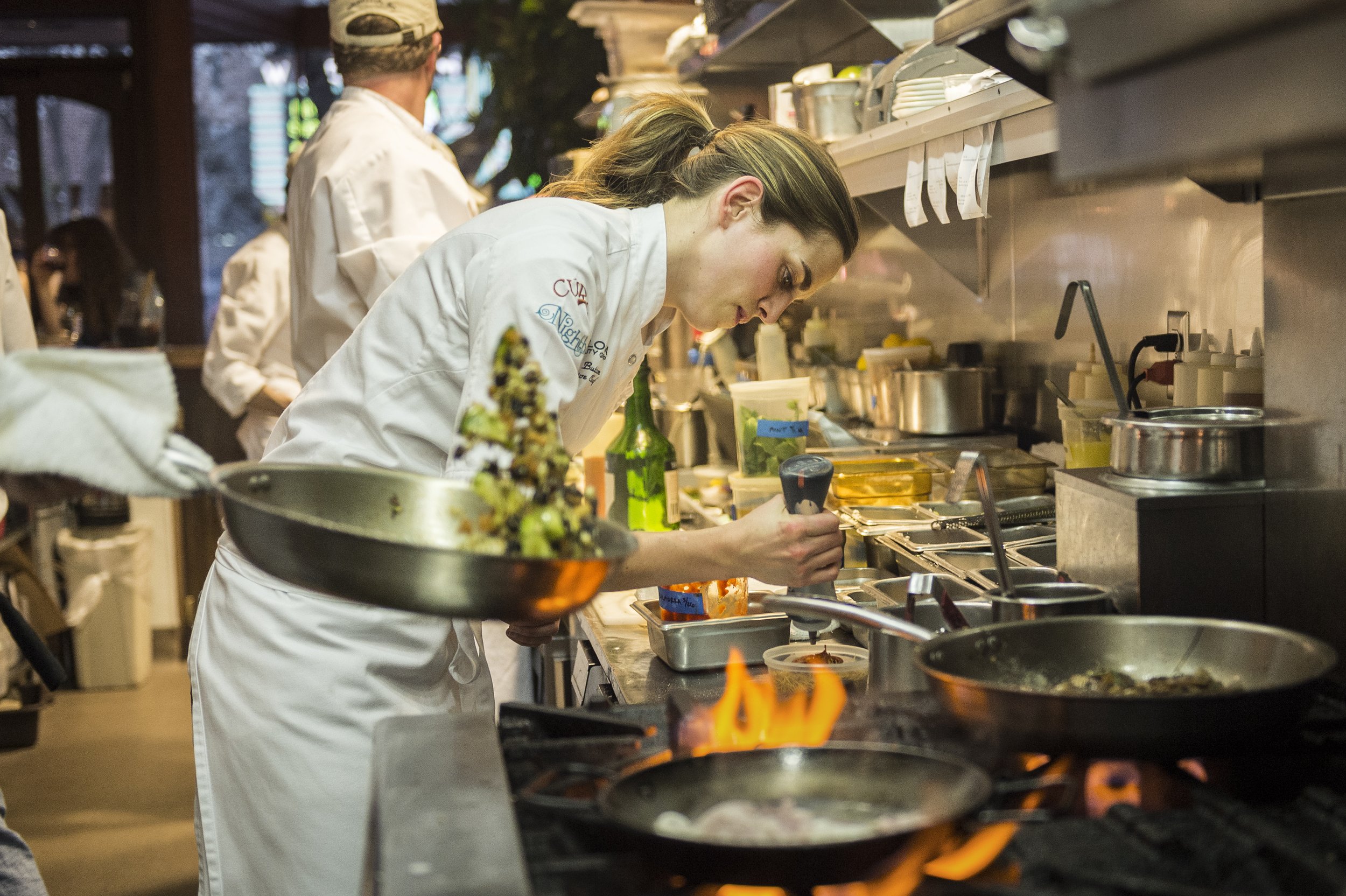 A female chef in a white uniform is cooking in a busy restaurant kitchen, holding a pan and focused on her work. Flames are visible from the stove, and there are various kitchen utensils and ingredients on the counter around her.