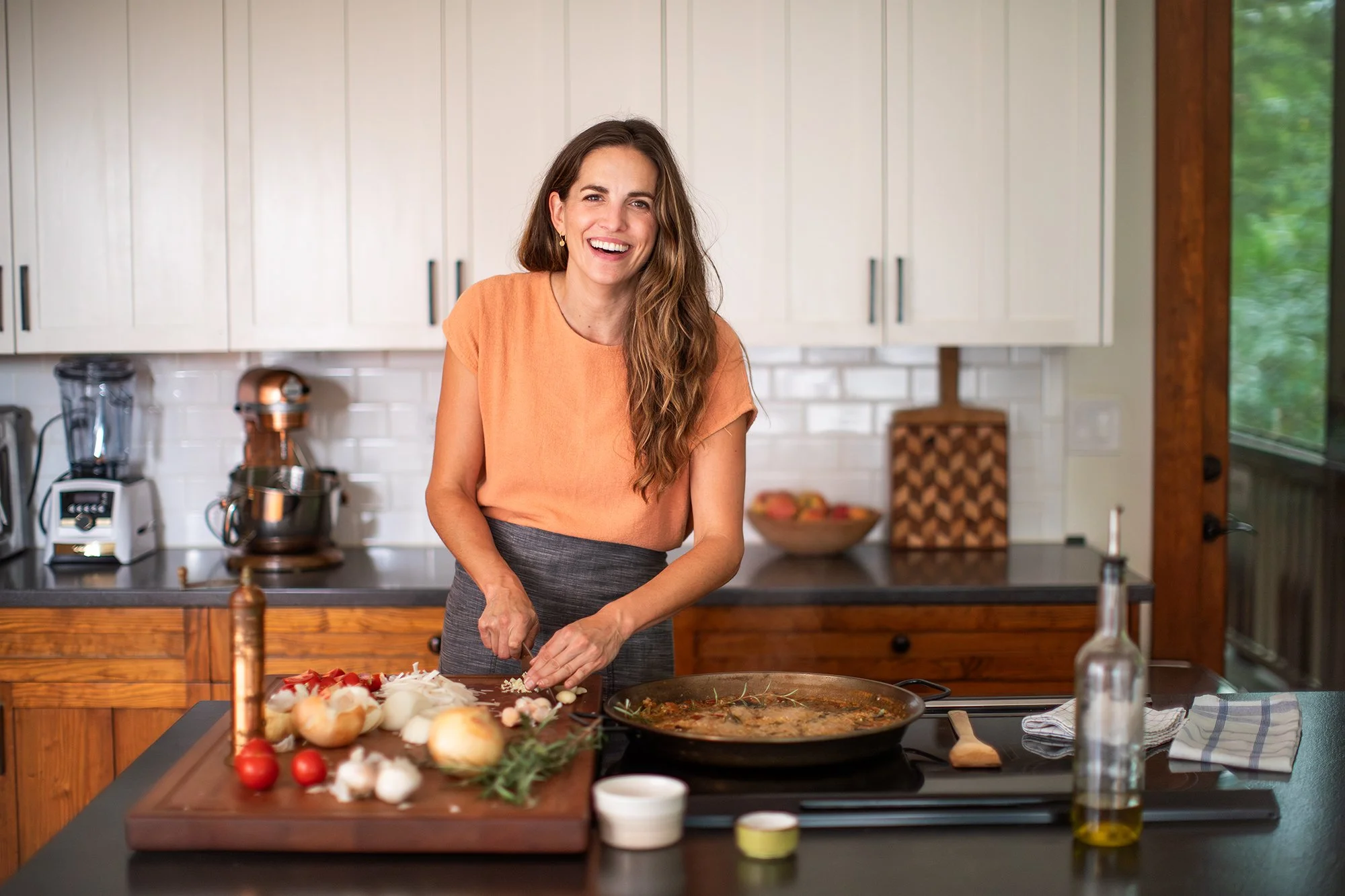 A woman in a kitchen chopping onions at a counter, with a pot of cooking food, olive oil, and cutting board with vegetables.