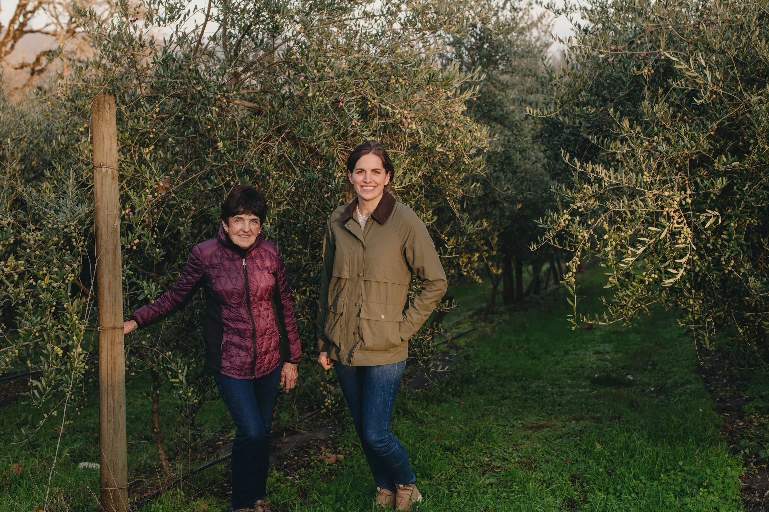 Two women standing together in an orchard surrounded by green trees and grass, smiling at the camera during daylight.