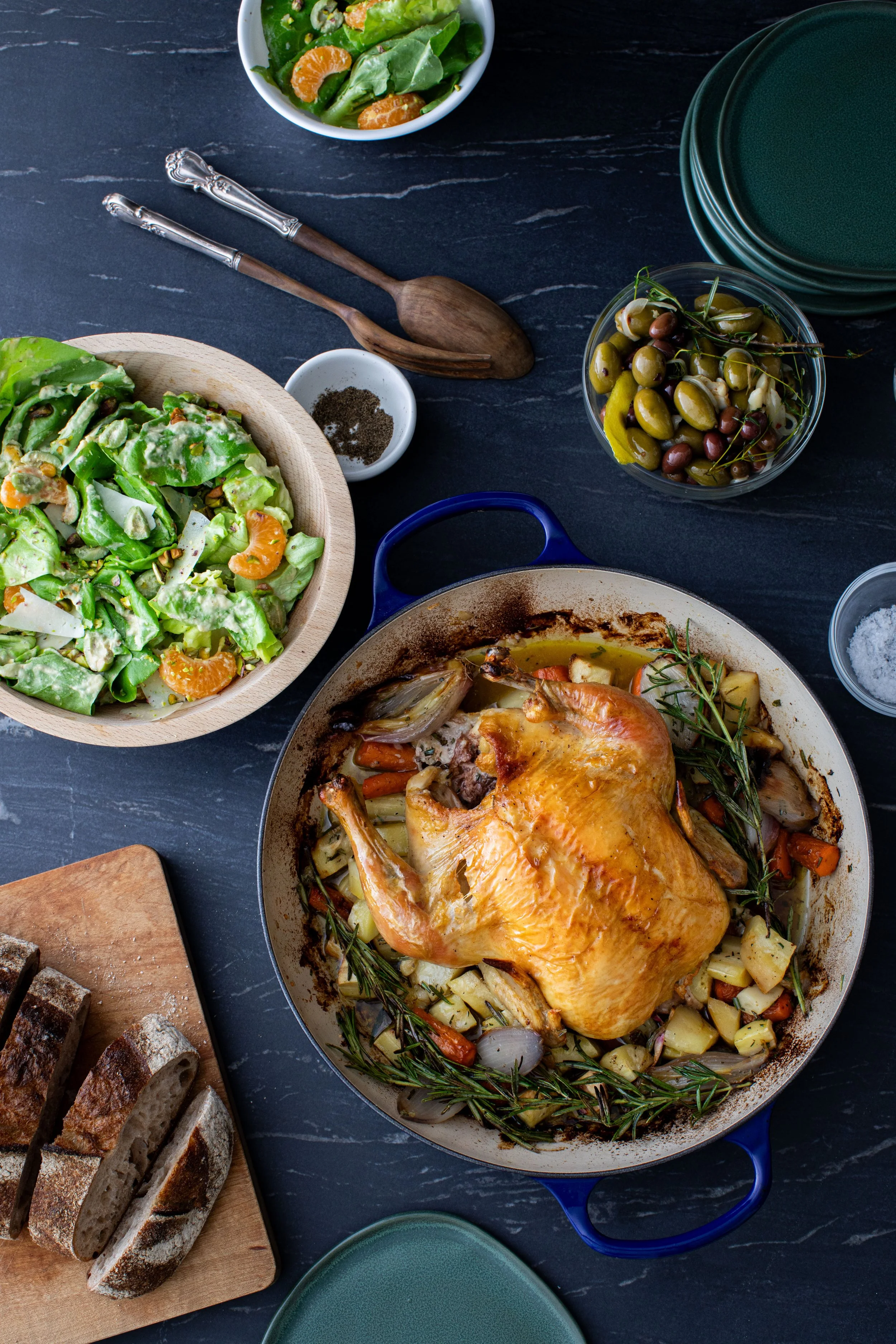 Roast chicken with roasted vegetables, green salad with mandarin slices, bowl of mixed olives, sliced bread, and empty plates on a dark table.
