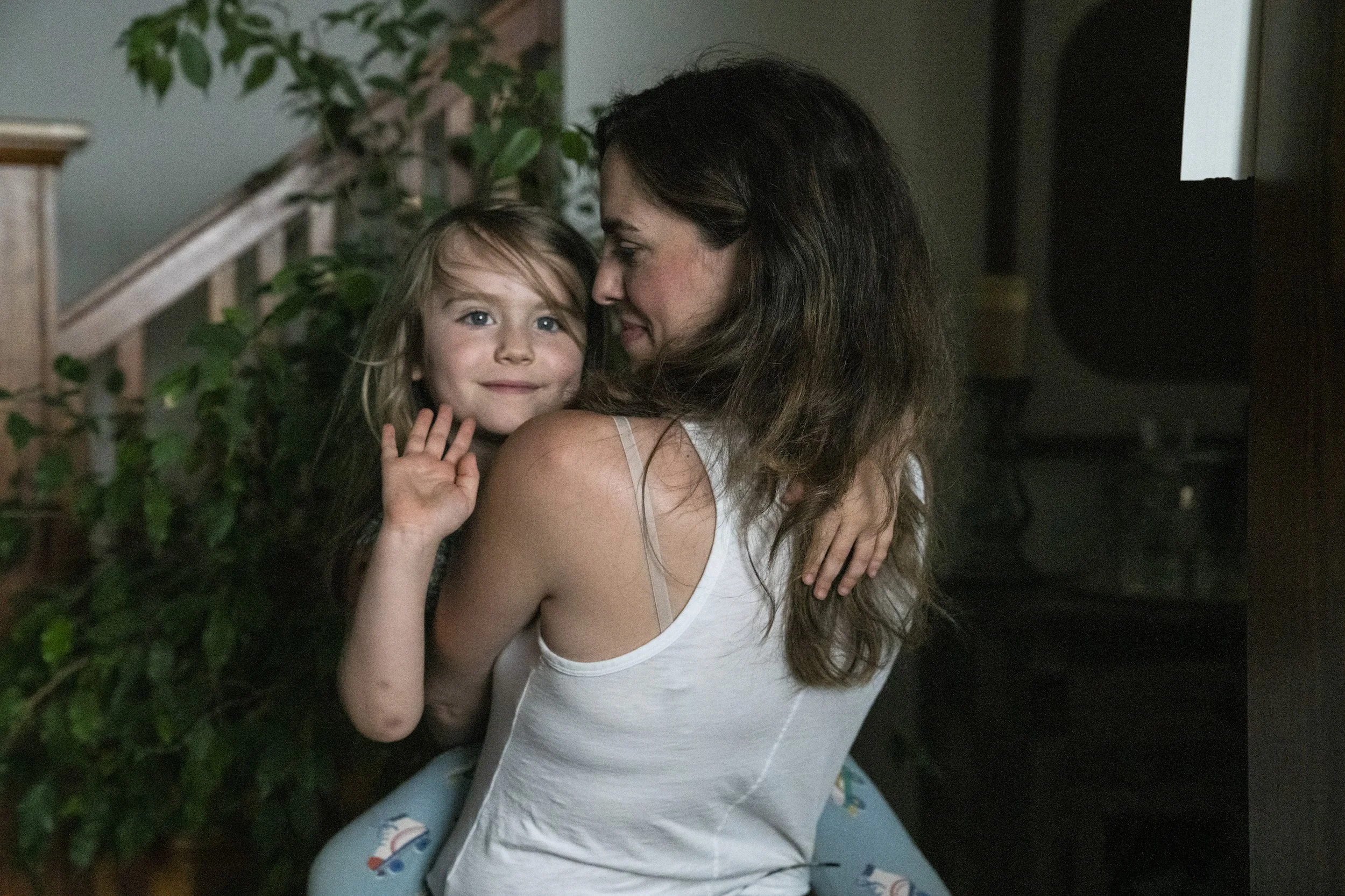 A woman embraces a young girl indoors, both smiling. The girl waves at the camera and has blonde hair and blue eyes. The woman has dark hair and is wearing a white tank top.