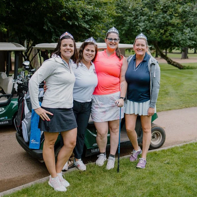 Four women standing on a golf course, wearing crowns, celebrating a golf event, with golf carts and trees in the background.