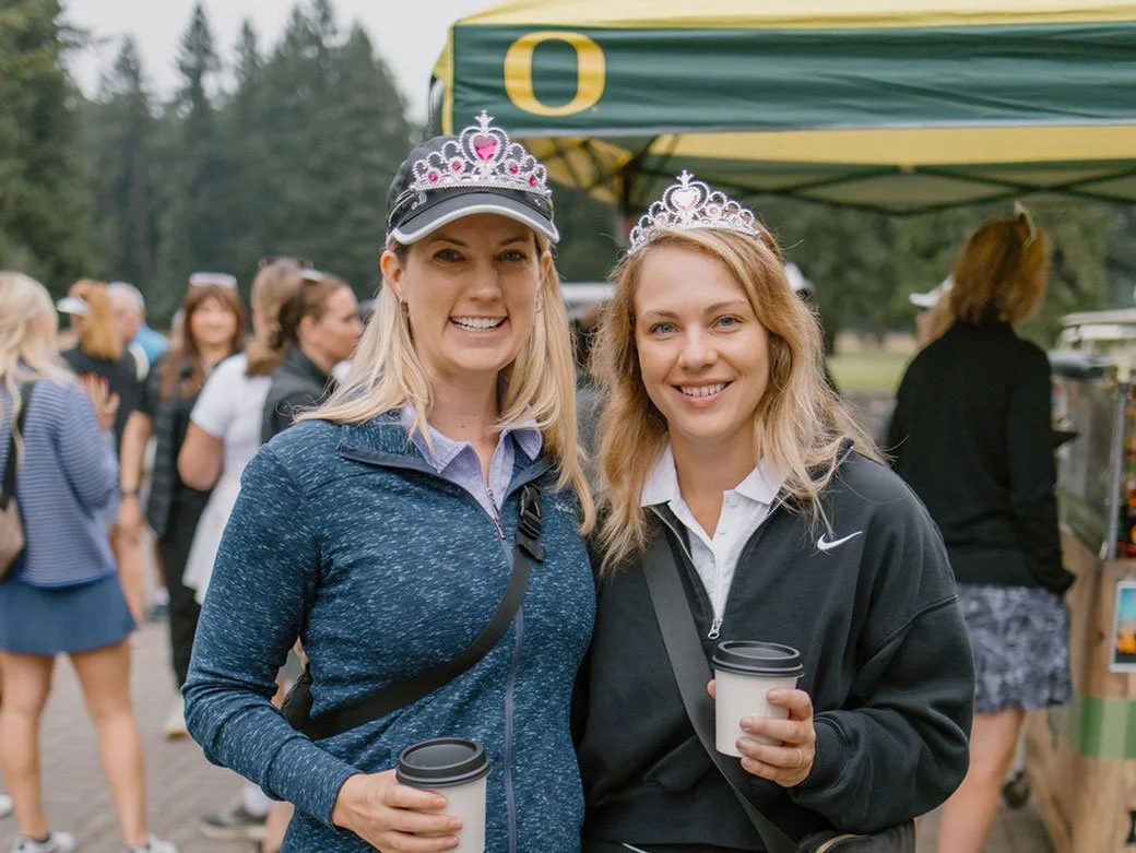 Two women wearing tiaras and holding coffee cups at an outdoor event, with a green and yellow tent and a group of people in the background.