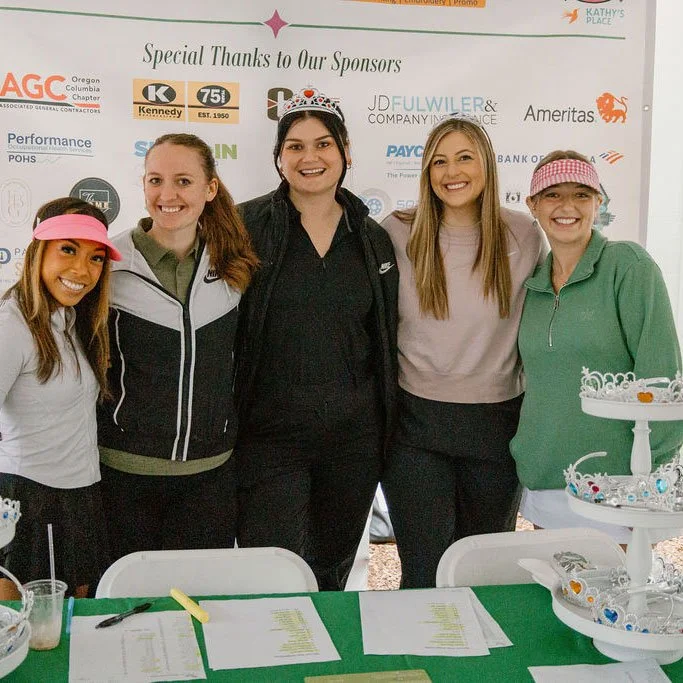 Five women stand together at a table during an event with a sponsor banner behind them. The women are smiling, and some are wearing casual sportswear or hats. The table has papers, pens, and a cake stand with decorated cupcakes.