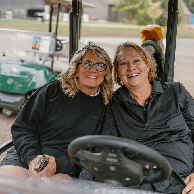 Two women sitting in a golf cart, smiling and posing for the photo with a golf course in the background.