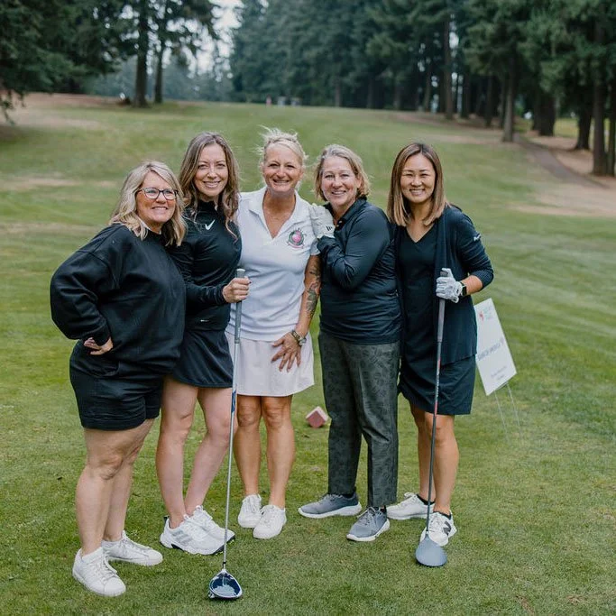 Five women standing on a golf course, smiling, holding golf clubs, dressed in casual golf attire, with trees and grass in the background.