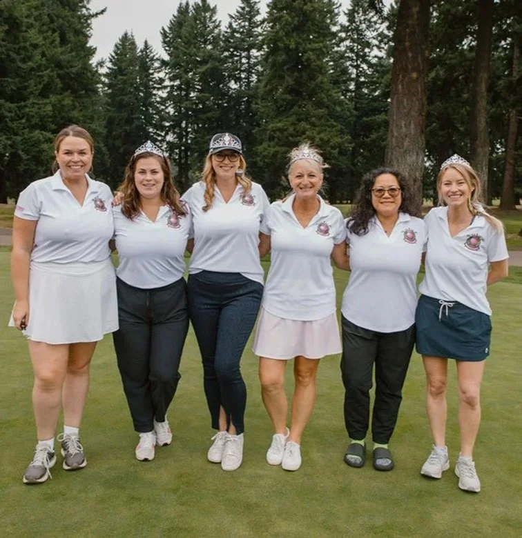 Six women standing on a golf course, some wearing tiaras and casual golf attire, with trees in the background.