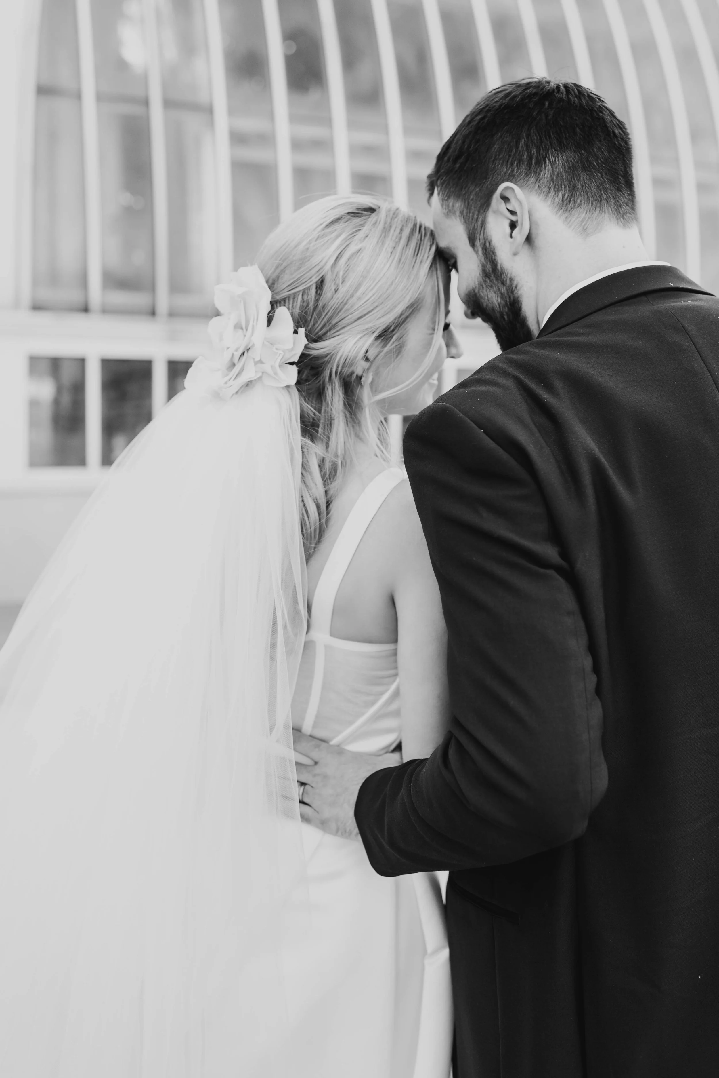 A bride and groom touch foreheads, while turned away from the camera. The bride is wearing an heirloom veil and the groom's hand is on her lower back. This photo is black and white.