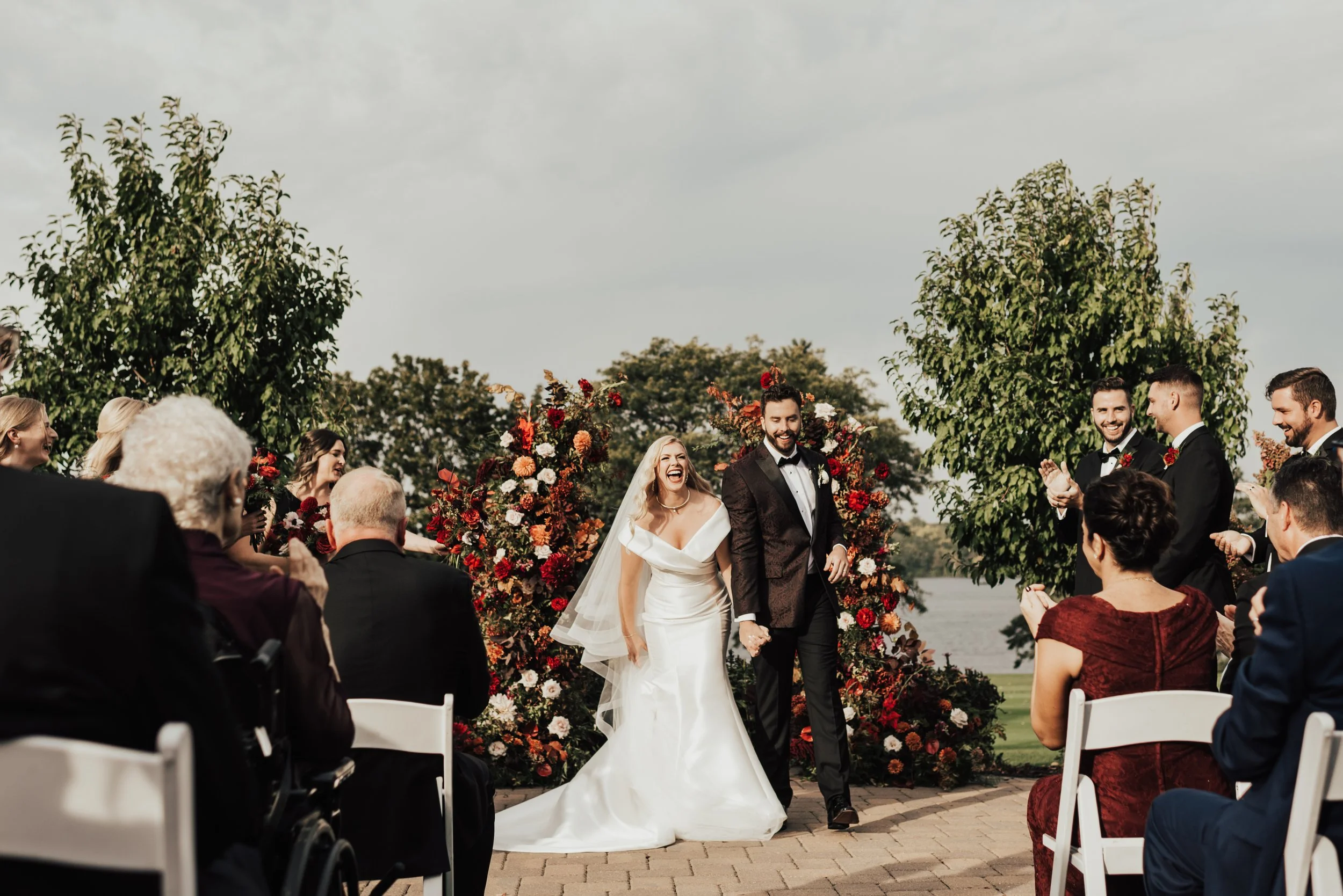 A bride and groom smile and laugh loudly after having their first kiss during their ceremony. They are standing outside by a lake, in front of their guests with a beautiful red and orange flower arch behind them.