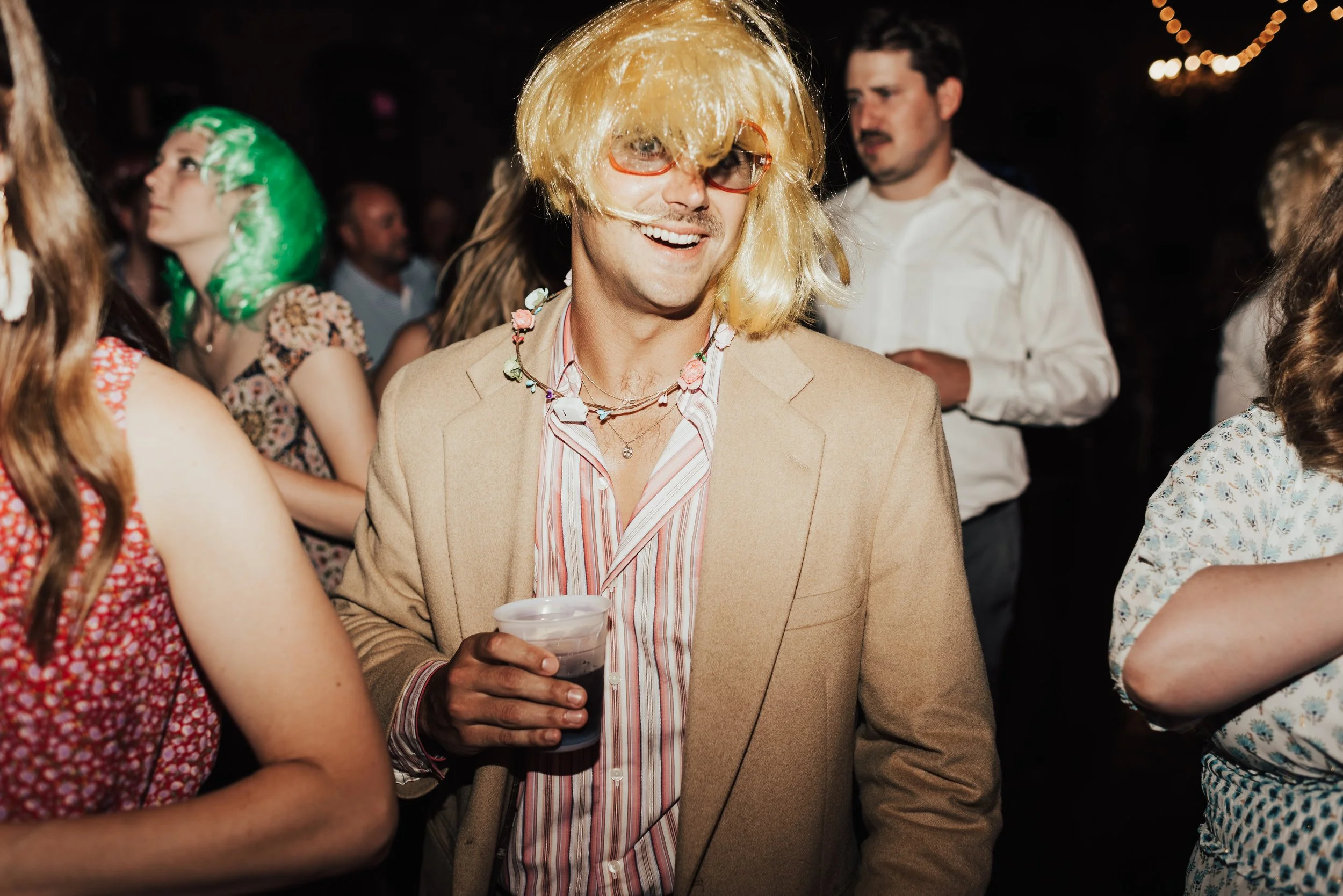 A wedding guest, wearing pink sunglasses and a blonde wig, holding a drink, is caught mid laugh in the middle of a dance floor.