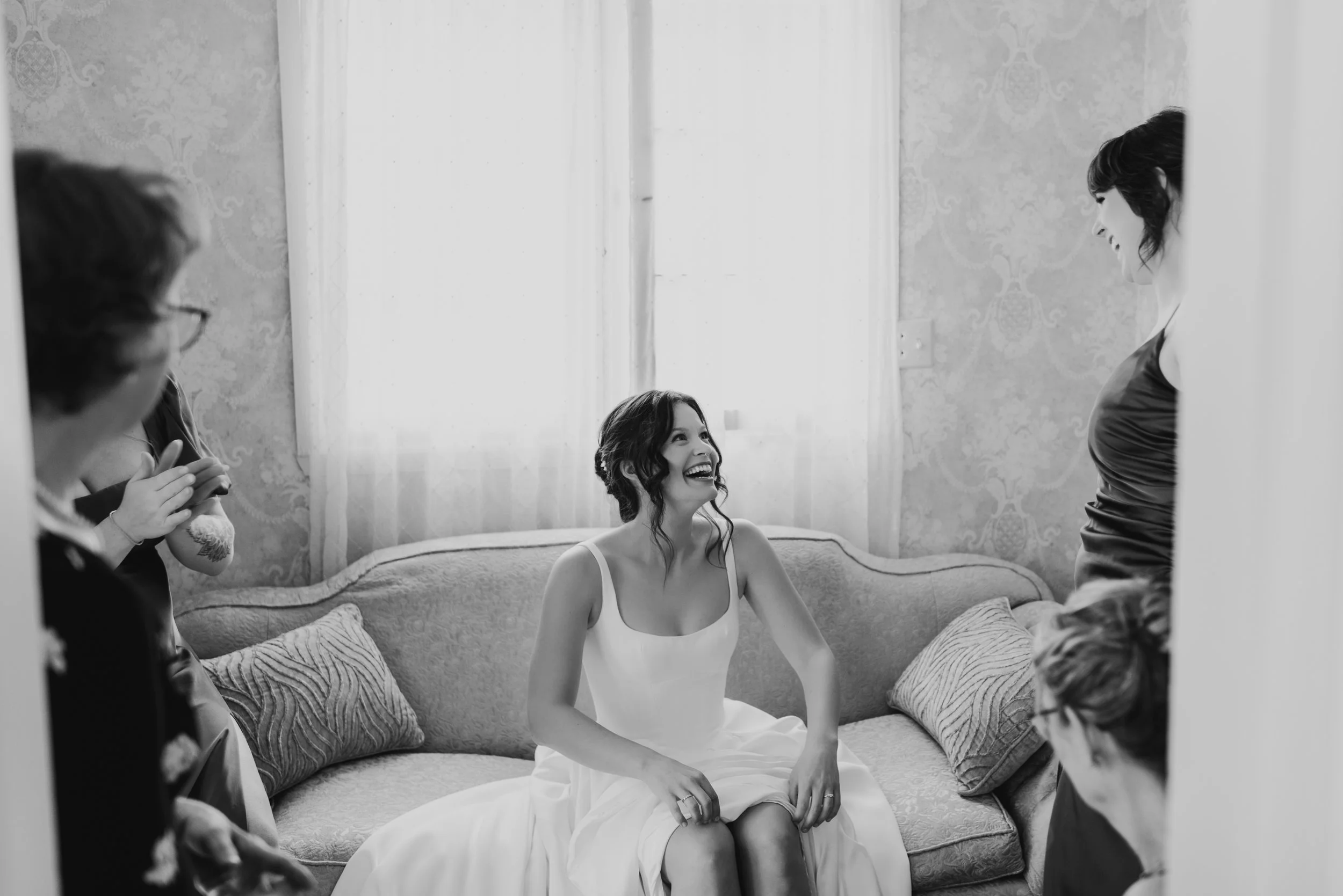 A candid black and white image of a bride sitting on a couch, surrounded by other women. She is laughing and looking up to her sister, who si standing to her left.