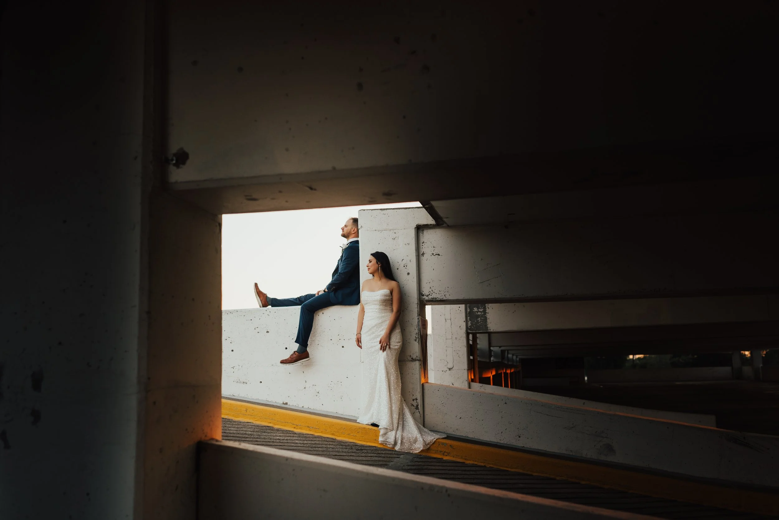 Bride and groom in wedding attire sitting on the edge of a parking garage ledge, with the groom sitting and looking out and the bride standing nearby, both appearing relaxed and contemplative.