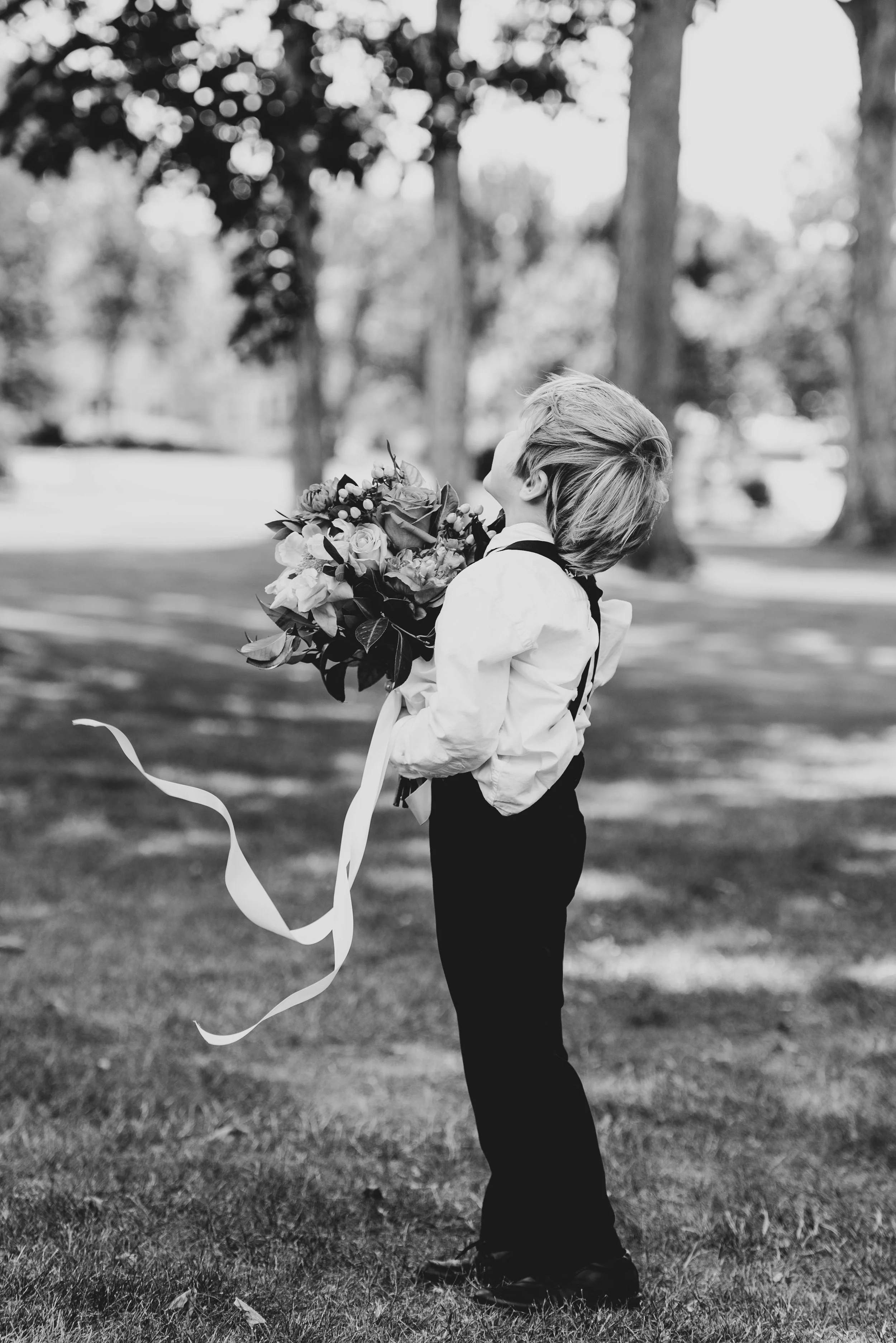 A little boy in wedding attire holds a brides bouquet. His back is turned and the ribbons are moving in the wind. This photo is black and white and was taken at Lafayette Club on Lake Minnetonka.