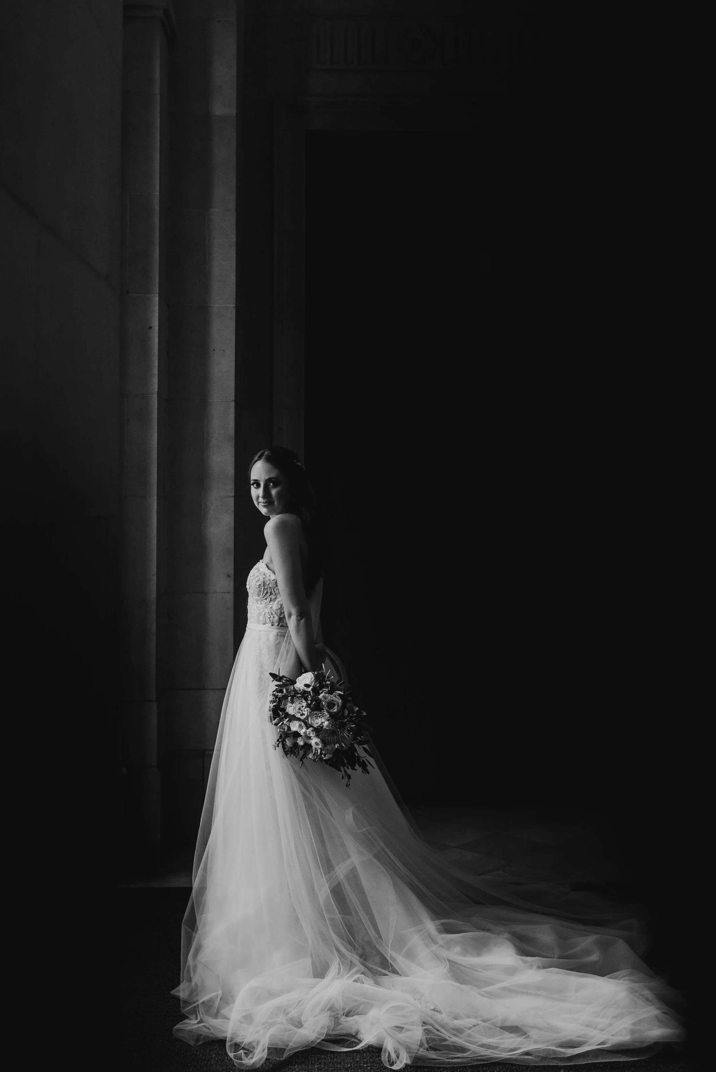 This is a moody black and white, fine art portrait of a bride. She is staring at the camera with her hands behind her back. This phot was taken at the Basilica St. Mary in Minneapolis, MN.