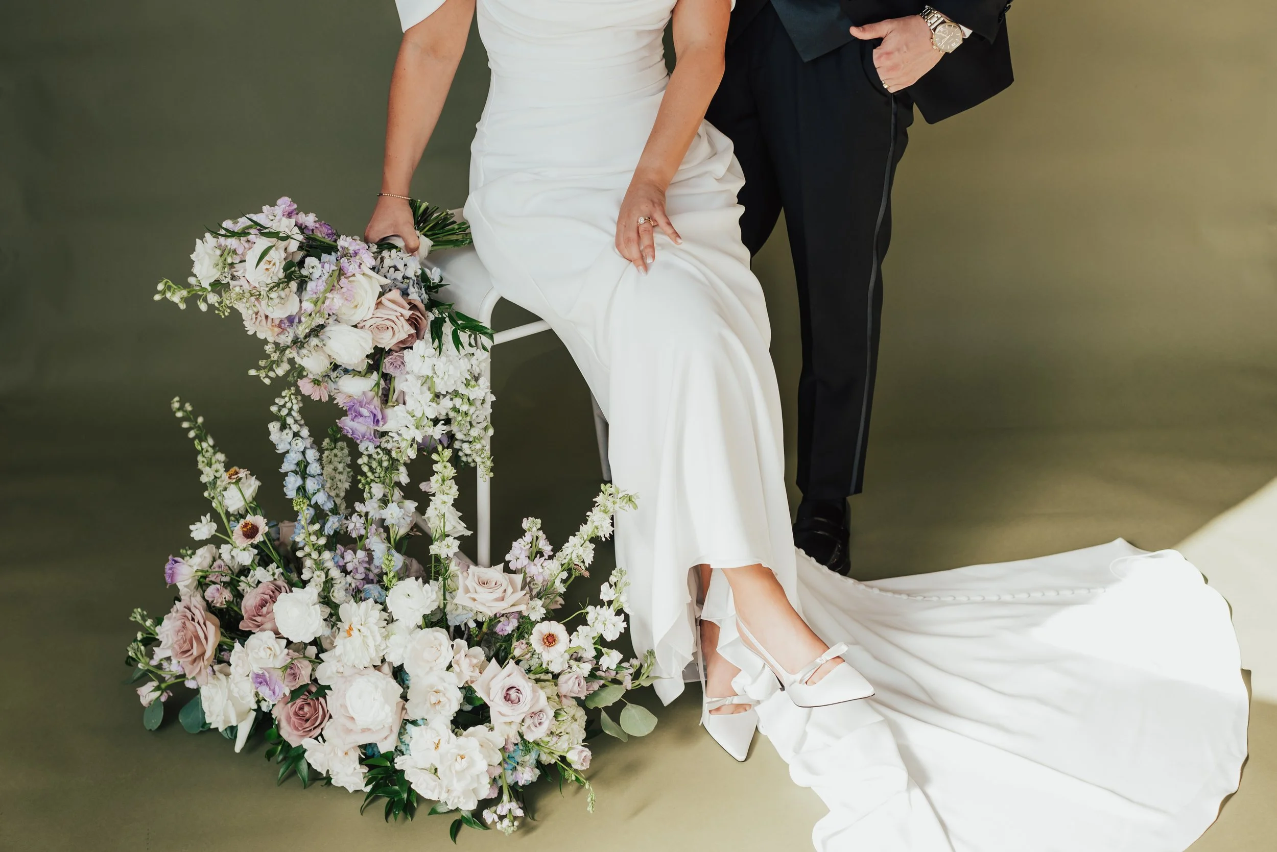 A bride sits on a white stool, showing her wedding shoes. The groom is standing beside her and they are both surrounded by flowers. This photo was taken against a green backdrop in a fine art studio.