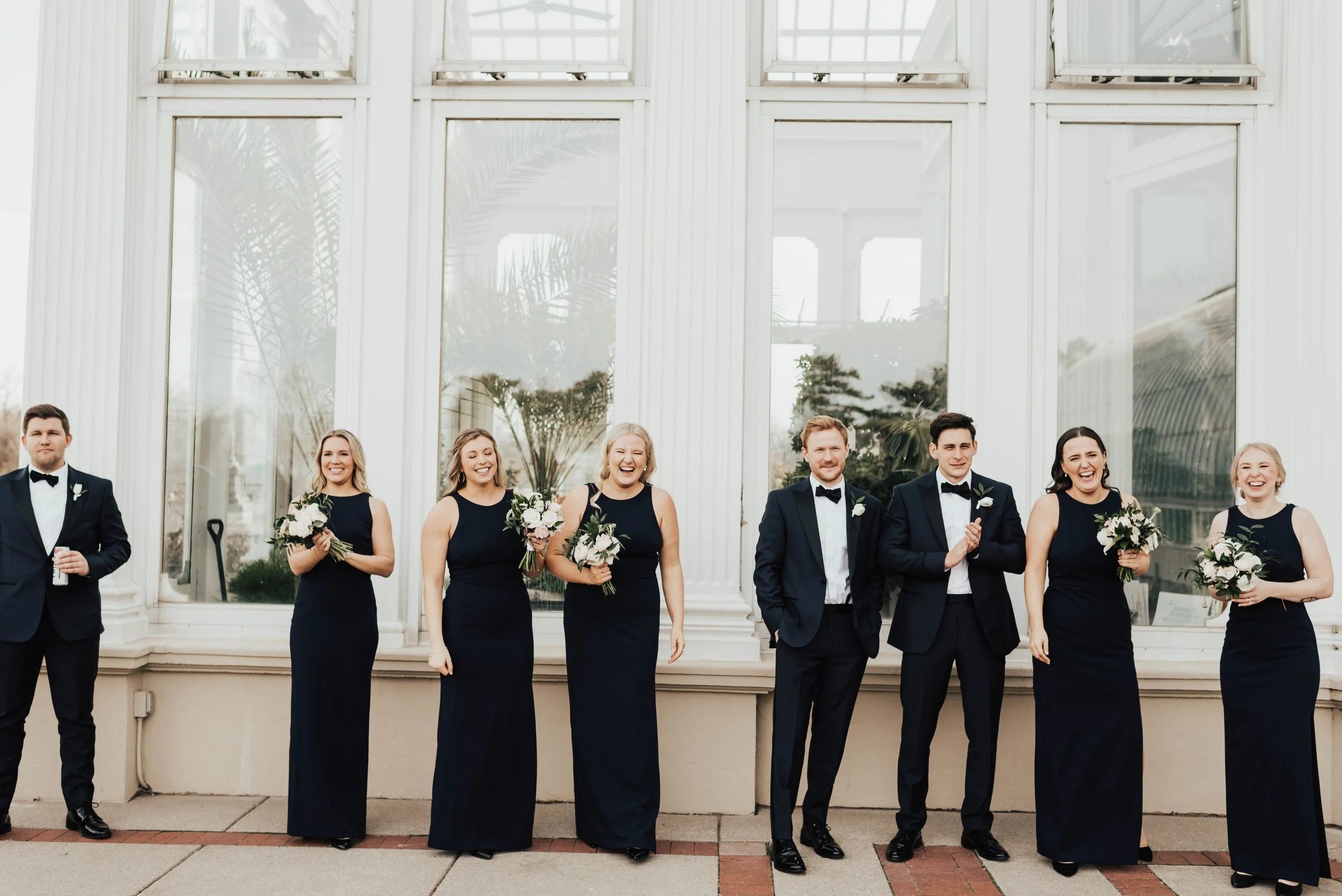 A line of bridal party members, dressed in tux's and black dresses holding bouquets laugh and talk to someone off camera. This photo was taken at the Como conservatory in St. Paul, MN.