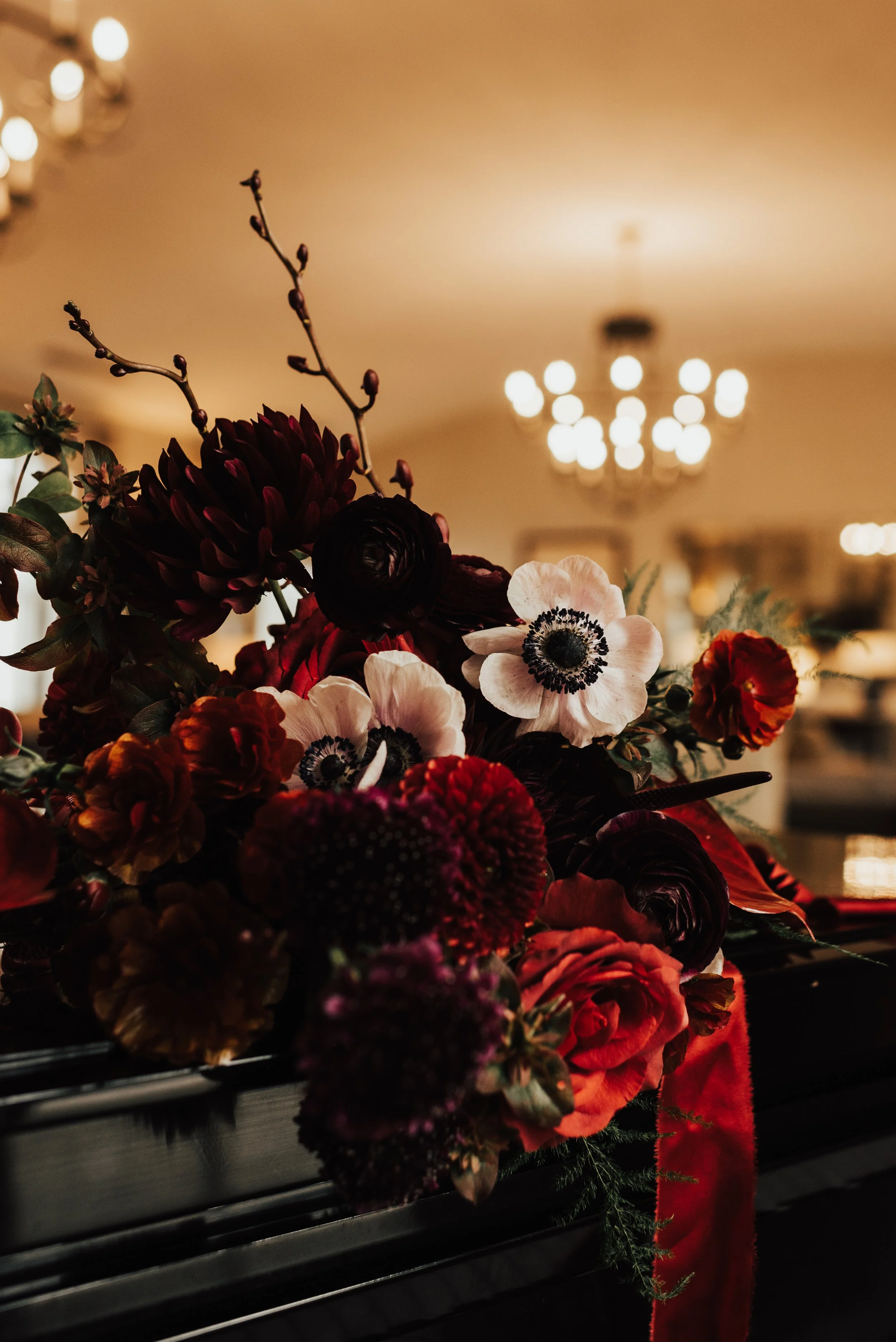 A deep red and light pink flowered bouquet is resting on a black piano, with soft warm light in the background.