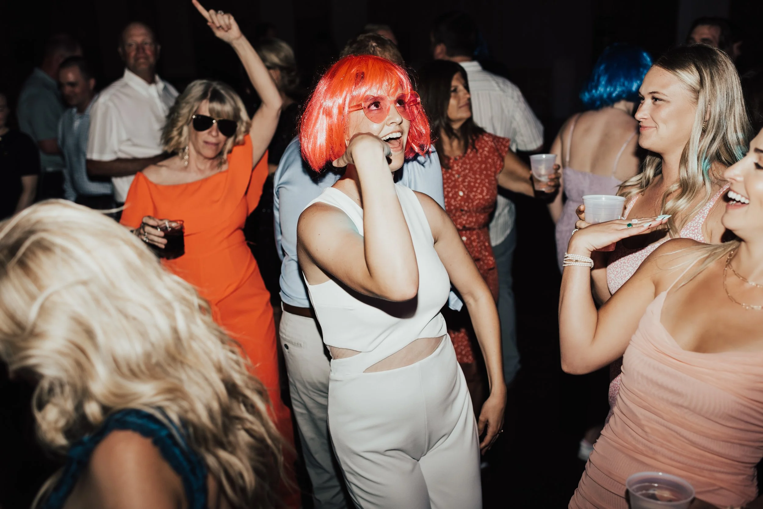 A bride in a white jumpsuit, lit in harsh flash, wearing a bright pink wig dances crazily with other girls.
