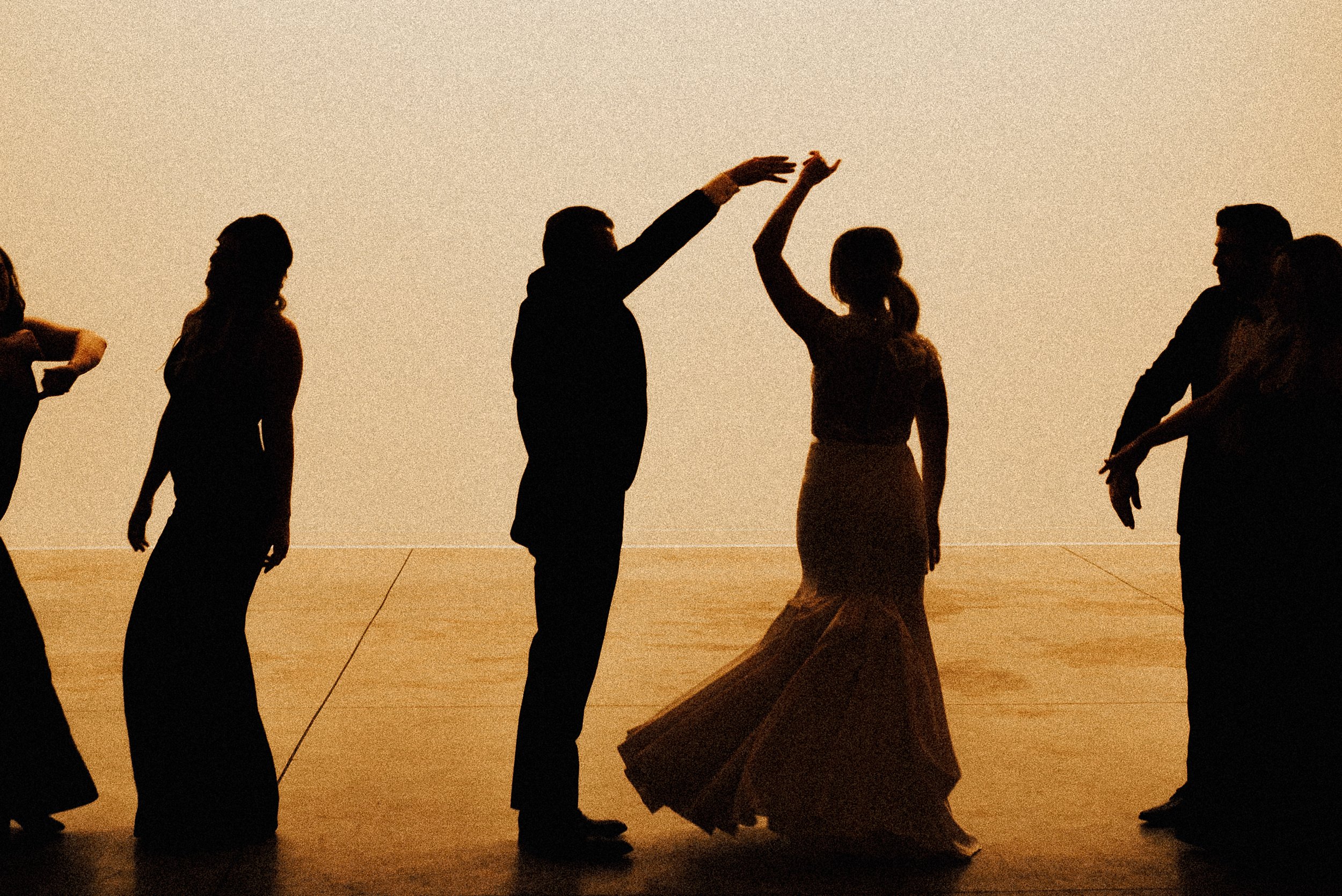 A photograph of people dancing. The people are sillouttes against a warm, grainy yellow backdrop and the bride + groom are centered. This photograph was taken at Pinewood in Cambridge, Minnesota.