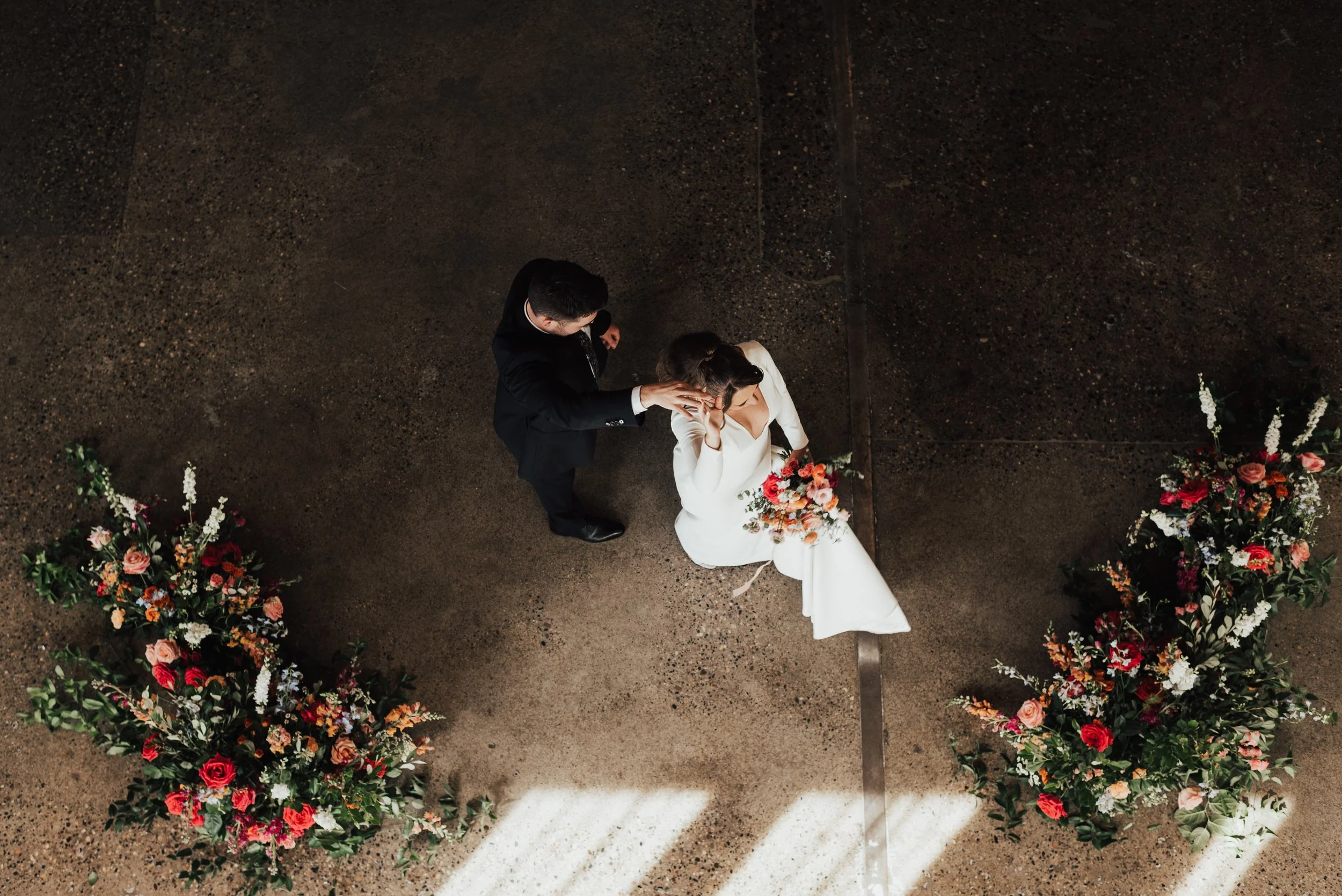 A groom is spinning a bride. This perspective is from above and they are winged by bright pink floral arrangements on either side. The bride carries a boquet with pink flowers.