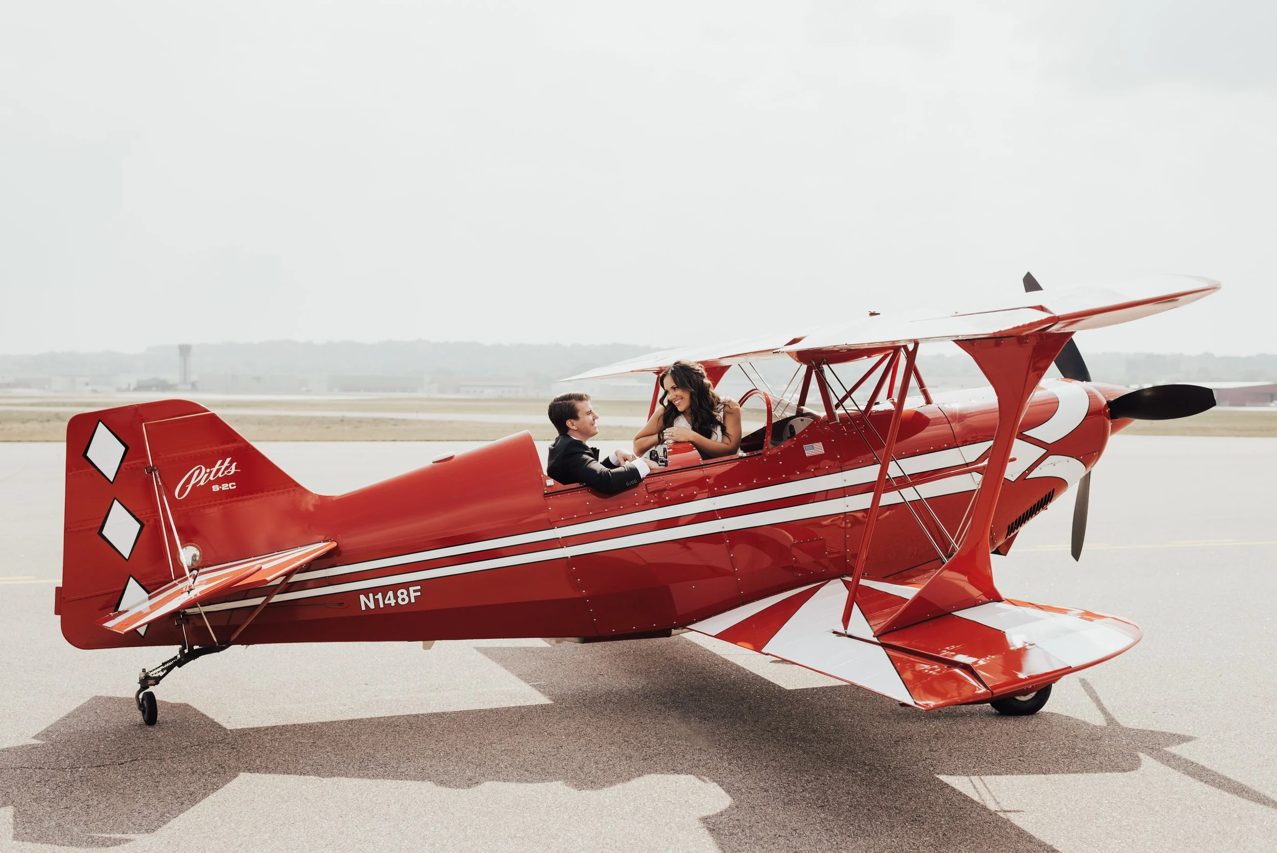 A bride is laughing at her groom while they both sit in a small, red airplane. The image was taken in full, bright sun in the middle of an airstrip in St. Paul, Minnesota.