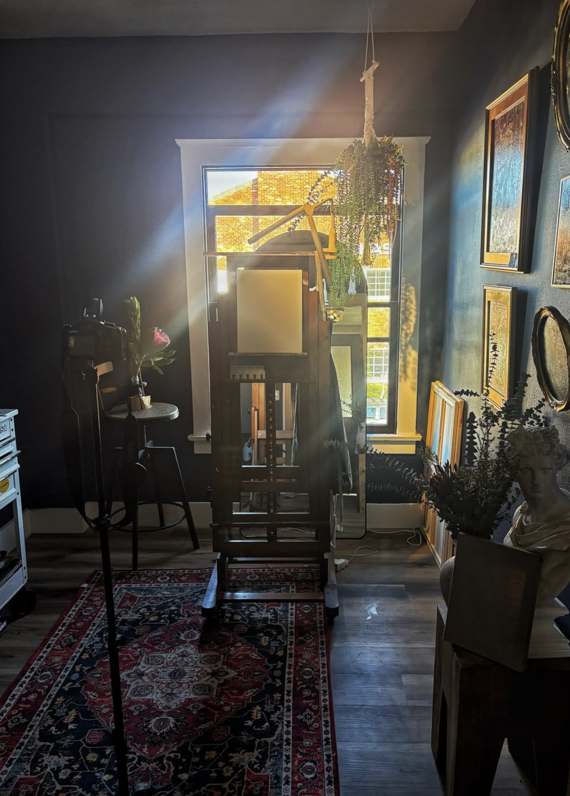 Sunlight coming through a window illuminating an artist's studio with art supplies, framed artwork, potted plants, and a bust sculpture.