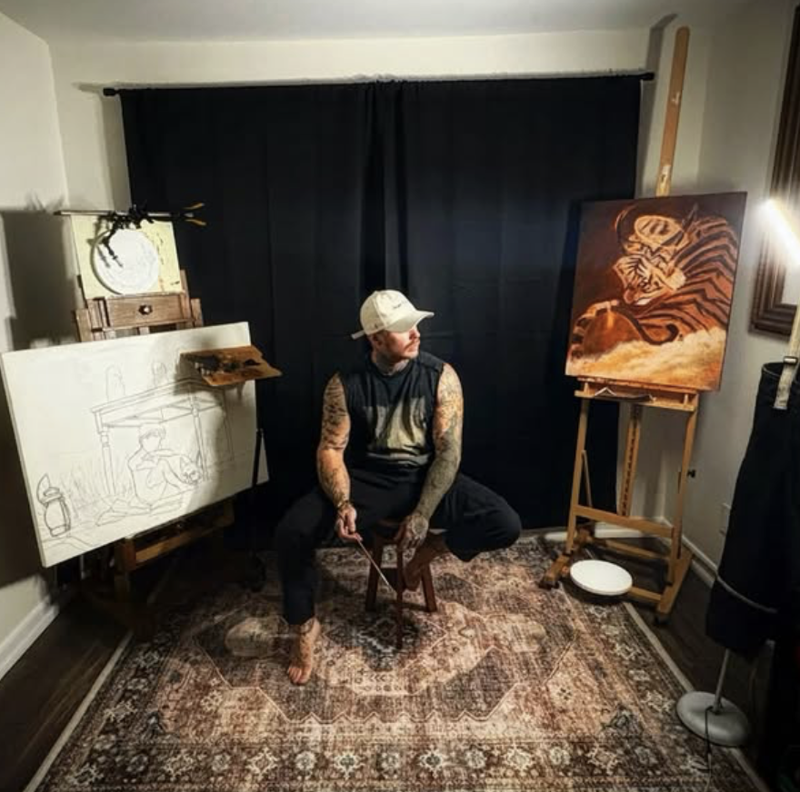 An artist sculpting in a small studio. The artist is seated on a stool, wearing a cap, sleeveless shirt, and black pants, with tattoos on his arms. The studio has a framed drawing on the left, a painting of a tiger on an easel to the right, and art supplies nearby. A Persian-style rug covers the floor.