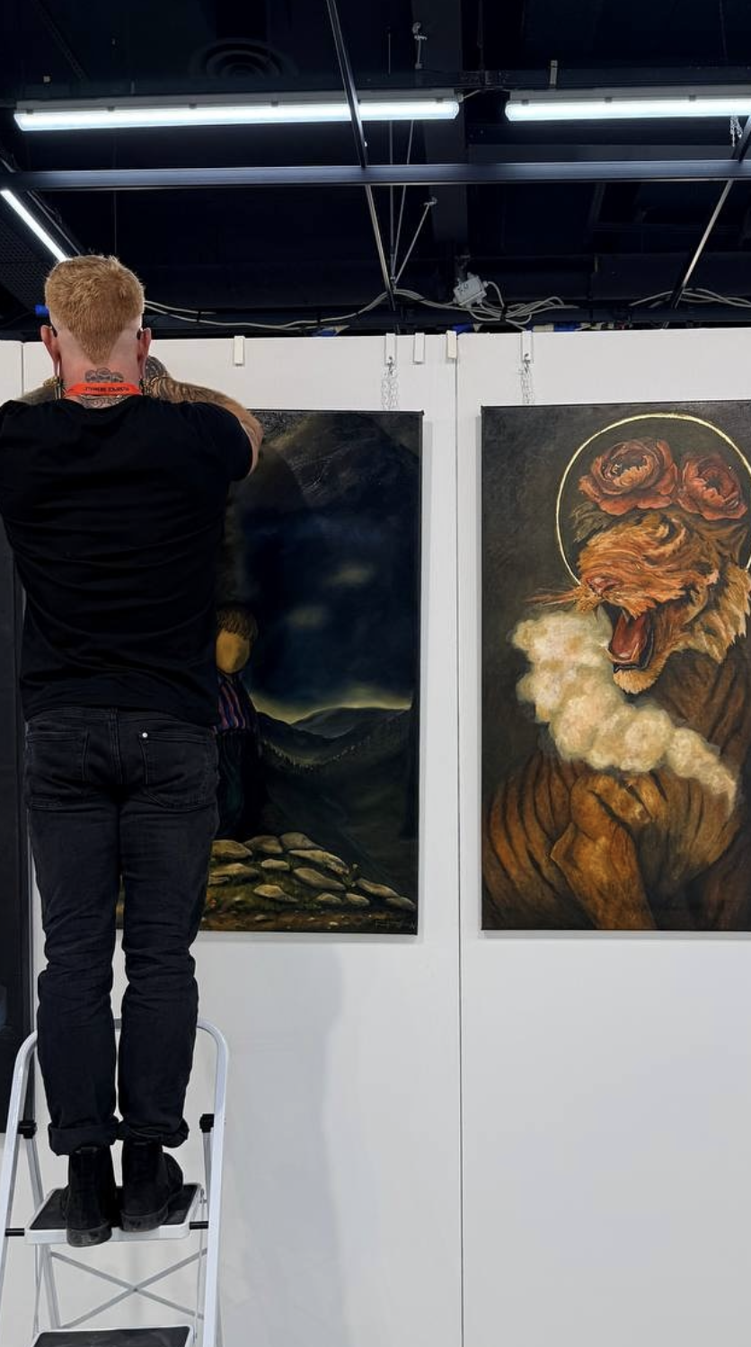 An artist hanging up paintings on a display wall at an art exhibition. The painting on the right depicts a roaring lion with a halo and a rose, with smoke emanating from its mouth. The painting on the left shows a dark landscape with mountains and a cloudy sky.