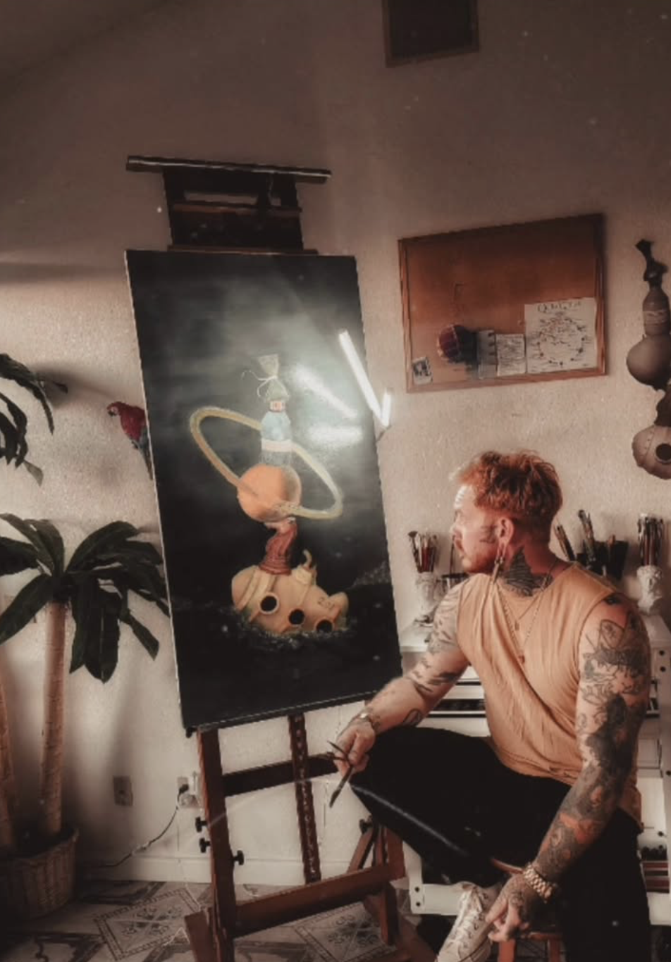 A man with tattoos, wearing a sleeveless shirt, sitting next to an easel with a canvas of an astronaut riding a rocket, inside a room with plants, a bulletin board, and art supplies.