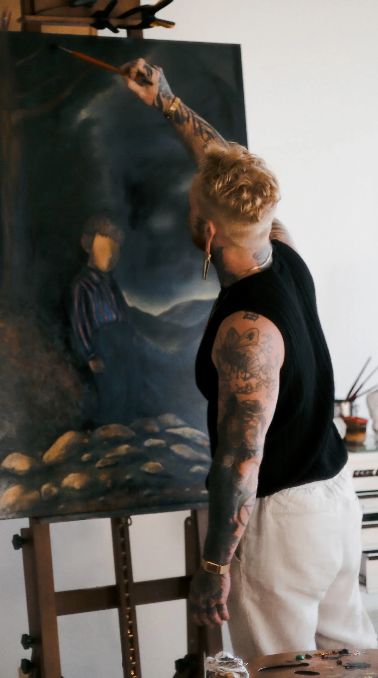 An artist with blonde hair, tattoos on arms, wearing a black sleeveless shirt and white pants, painting a portrait on a large canvas in a studio.