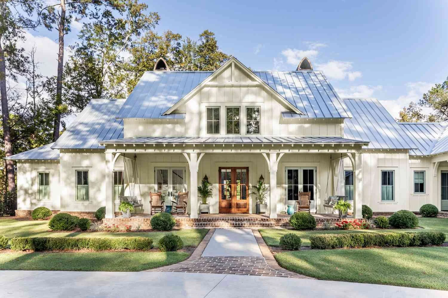 A white farmhouse-style house with a metal roof, front porch with swing chairs, potted plants, and brick steps leading to the wooden front door. Well-maintained green lawn and shrubbery surround the house, with tall trees in the background and a partly cloudy sky overhead.
