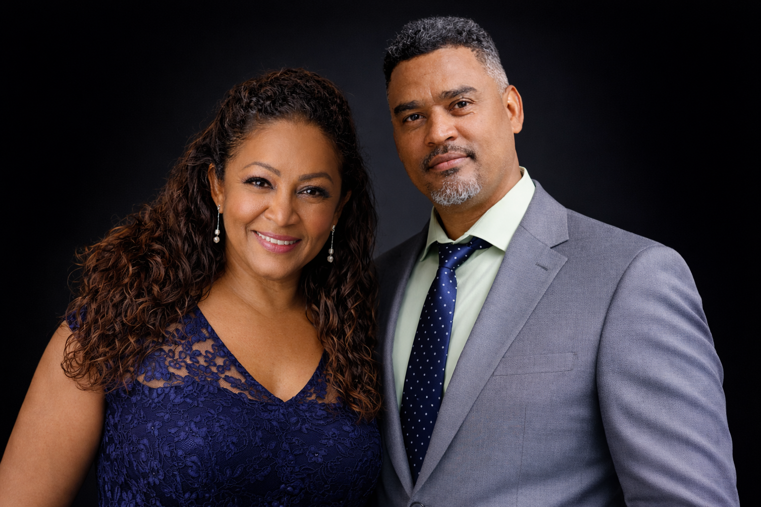 A middle-aged couple dressed in formal attire, posing together against a black background. The woman is wearing a navy blue lace dress with pearl earrings, and the man is in a light gray suit with a pale green shirt and a navy blue tie with white dots.