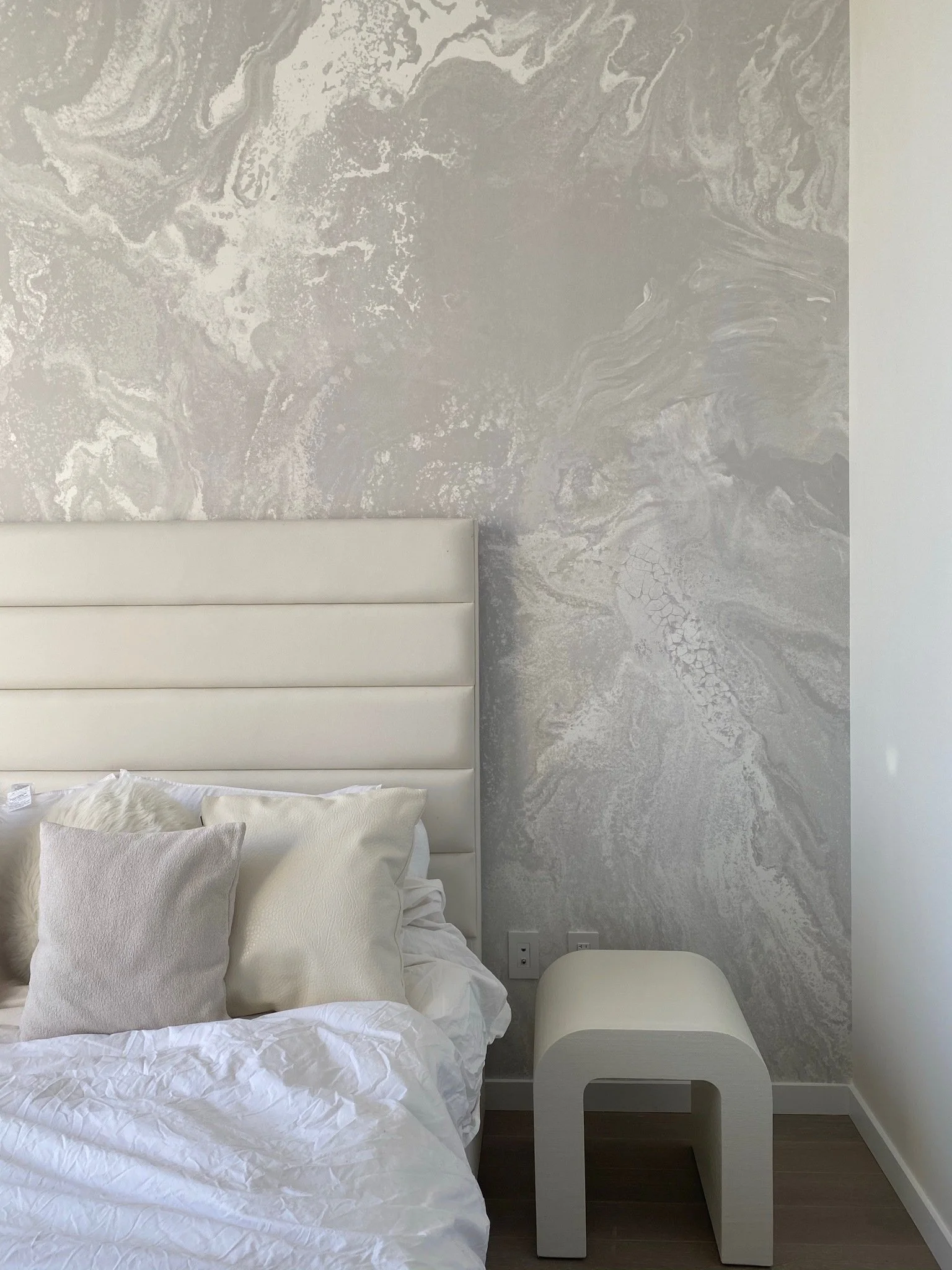 A bedroom with a white, padded headboard, white pillows, and white bedding. A small, white, modern side table with a curved design is beside the bed. The wall behind the bed has a gray and white marbled pattern.