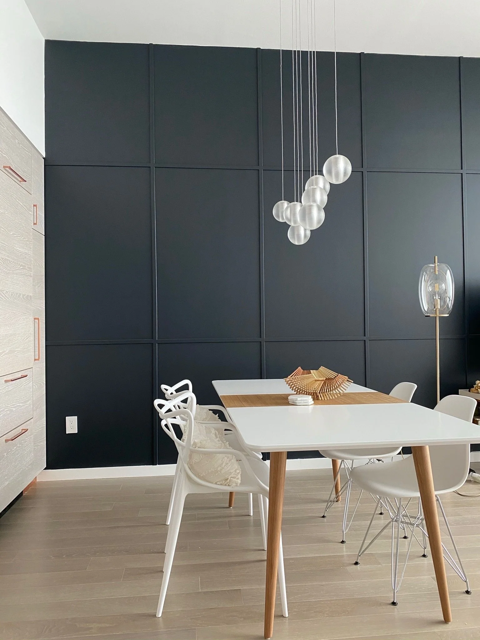 Modern dining room with a white table and white chairs, including a unique white chair with a cushion, against a dark paneled accent wall with pendant lighting and a wooden sideboard.