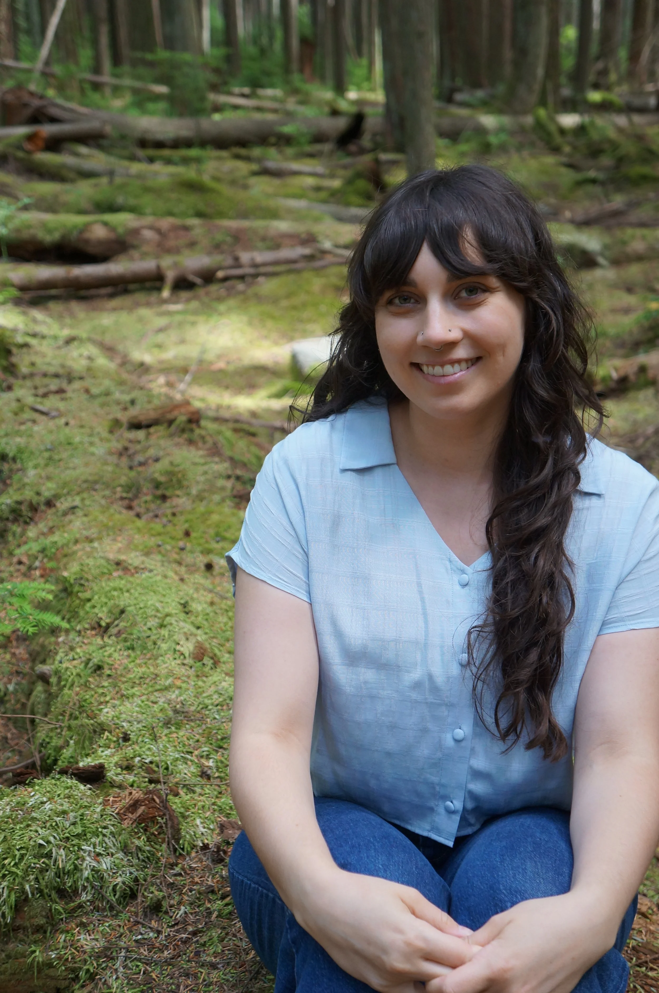 A woman with dark wavy hair and a septum piercing smiling while sitting in a forest with green moss and trees in the background.