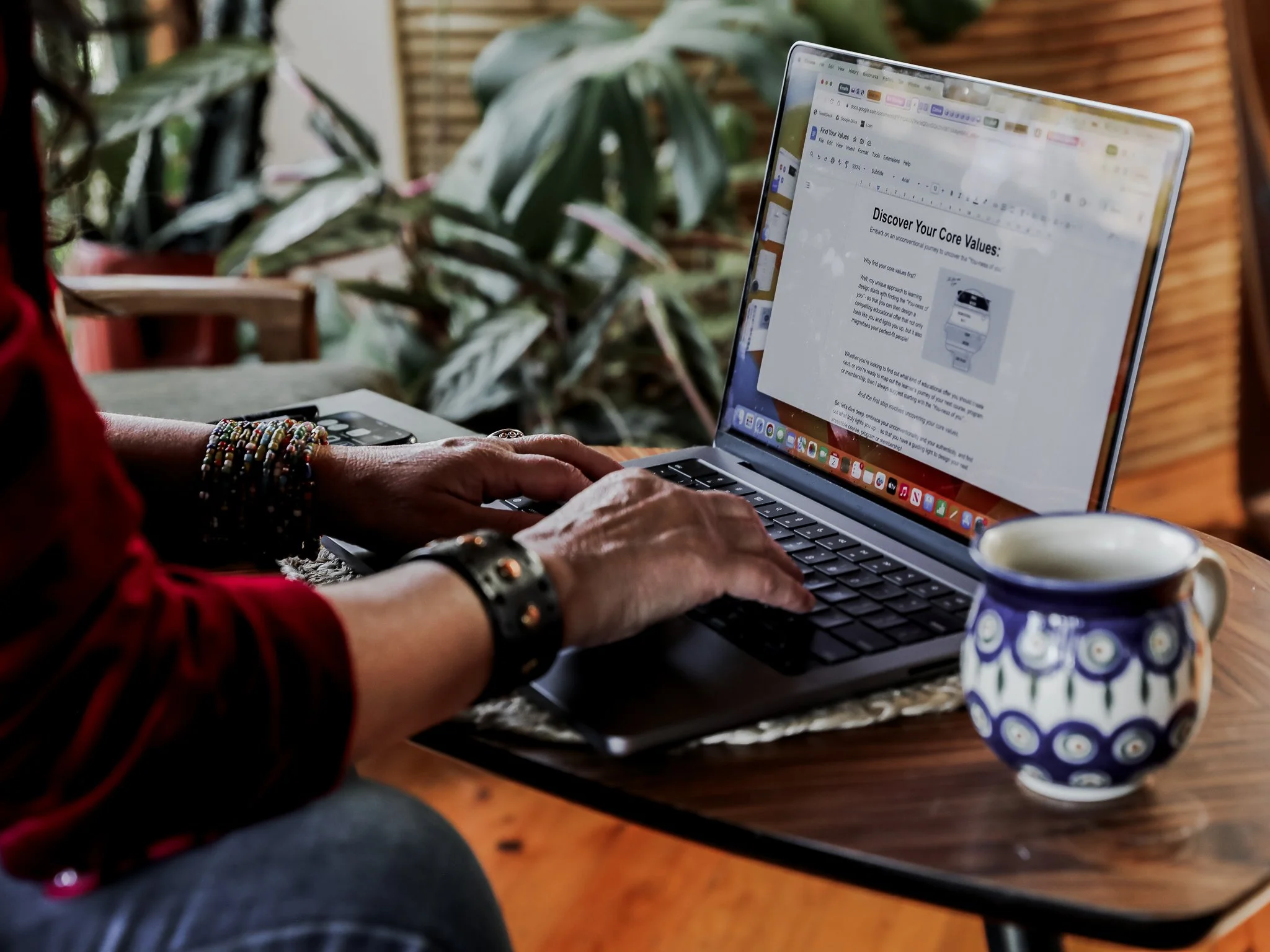 Tiffany Sinton working on a webpage on her laptop, sitting at a wooden table with a cup of coffee, and lush green plants in the background.