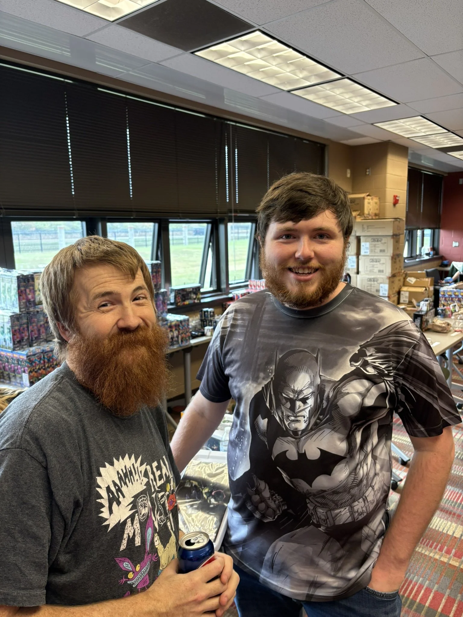 Two men with beards smiling indoors at a table with collectibles; one wearing a graphic t-shirt of Batman, the other in a T-shirt with a cartoon graphic and holding a soda can.