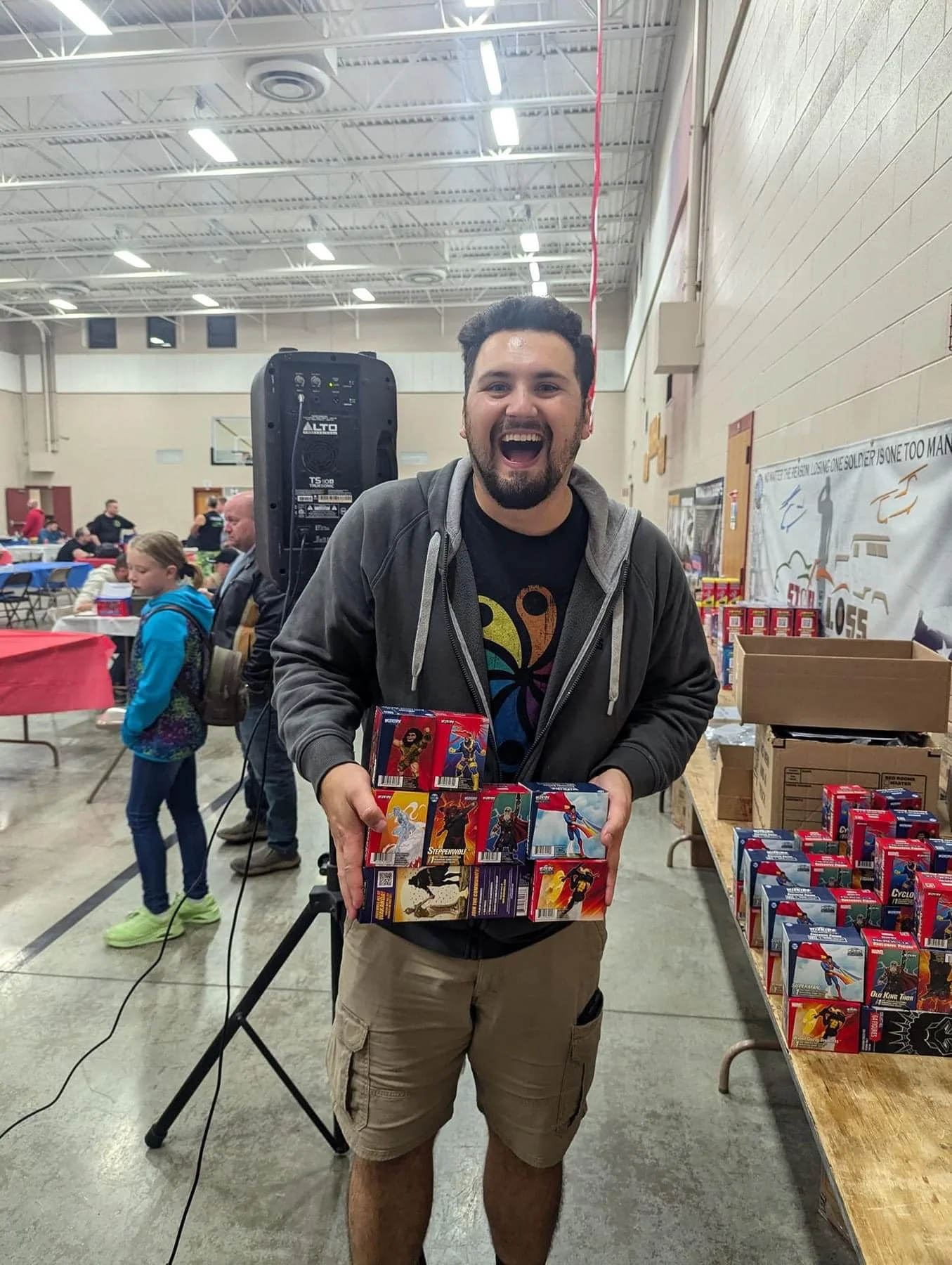 A man smiling and holding a collection of colorful boxes of toys or figurines, standing in a community center or gymnasium with tables and other people in the background.