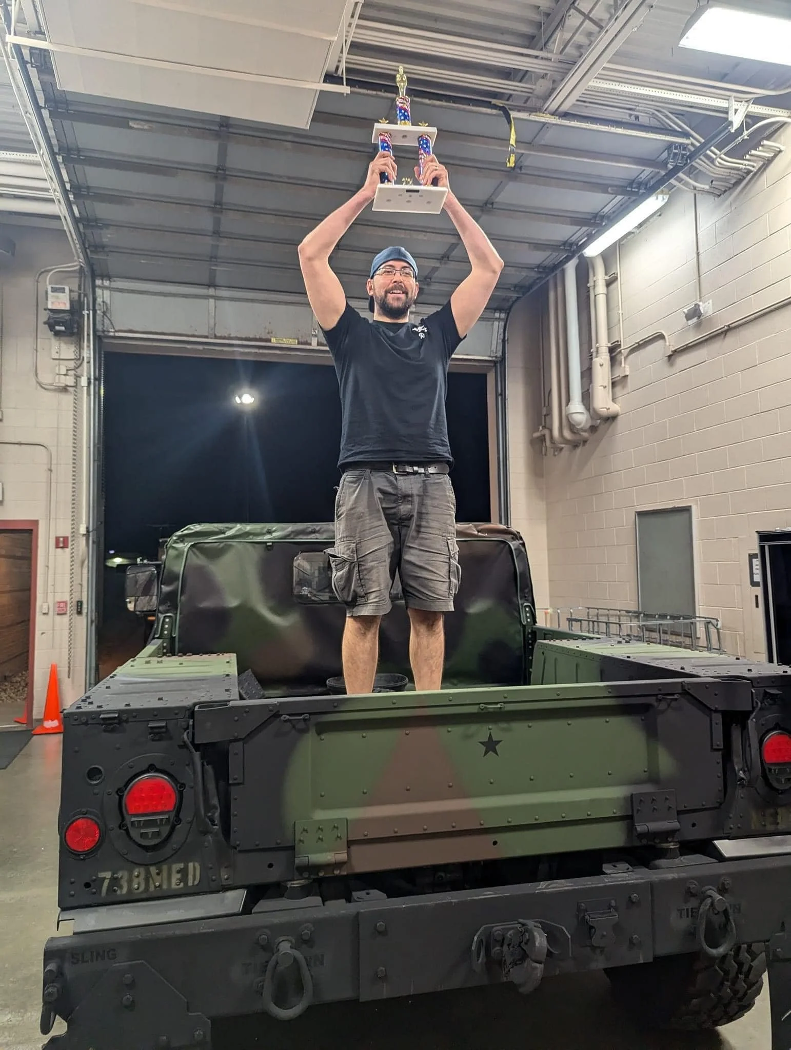 A man standing in the back of a military truck, holding a trophy above his head and smiling. The truck is in a warehouse or garage with an open door behind him.