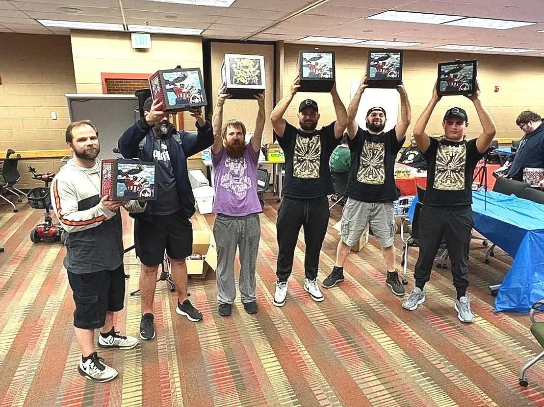 Six men standing side by side in a room, holding up boxes above their heads. The room has beige walls, a carpeted floor with a striped pattern, and tables covered with blue and yellow tablecloths. Other people are visible in the background.