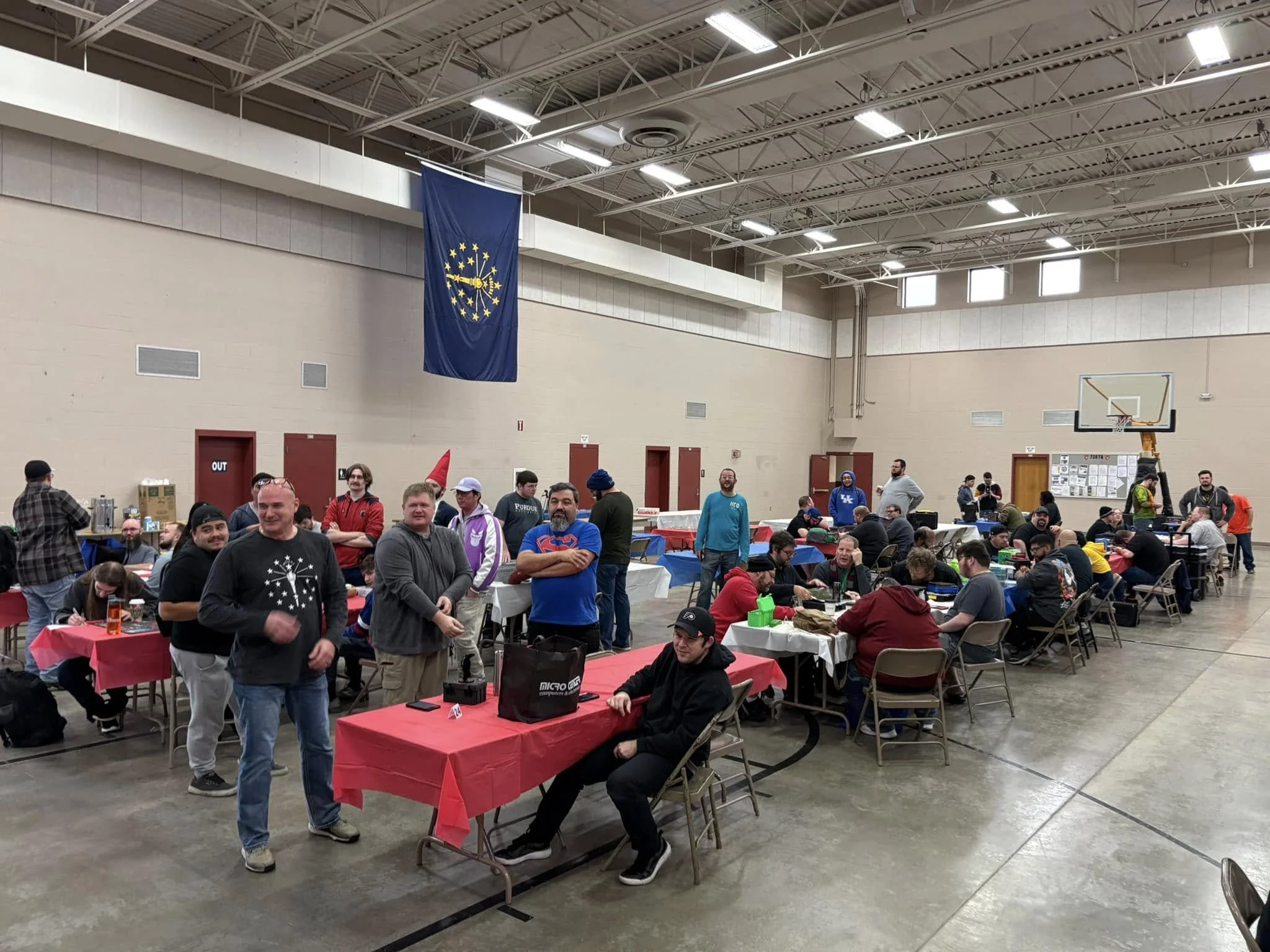 People gathered in a gymnasium with tables and chairs, some standing and some seated, engaging in social activities. A large Indiana state flag hangs from the ceiling, and the gym has a basketball hoop and high ceiling with bright lighting.
