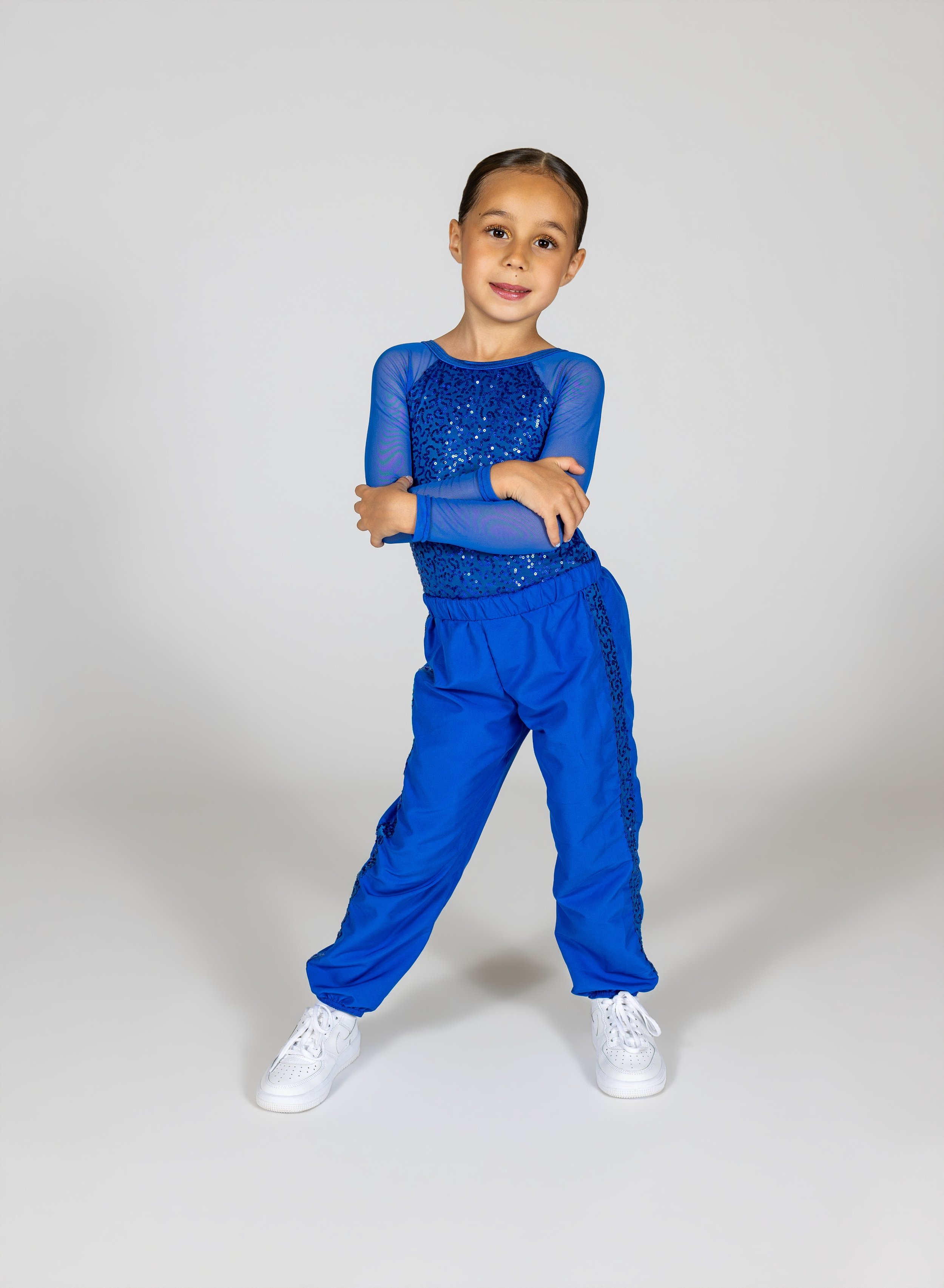 Dancer in a  blue dance outfit with sequins, white sneakers, standing with crossed arms, smiling, against gray background.