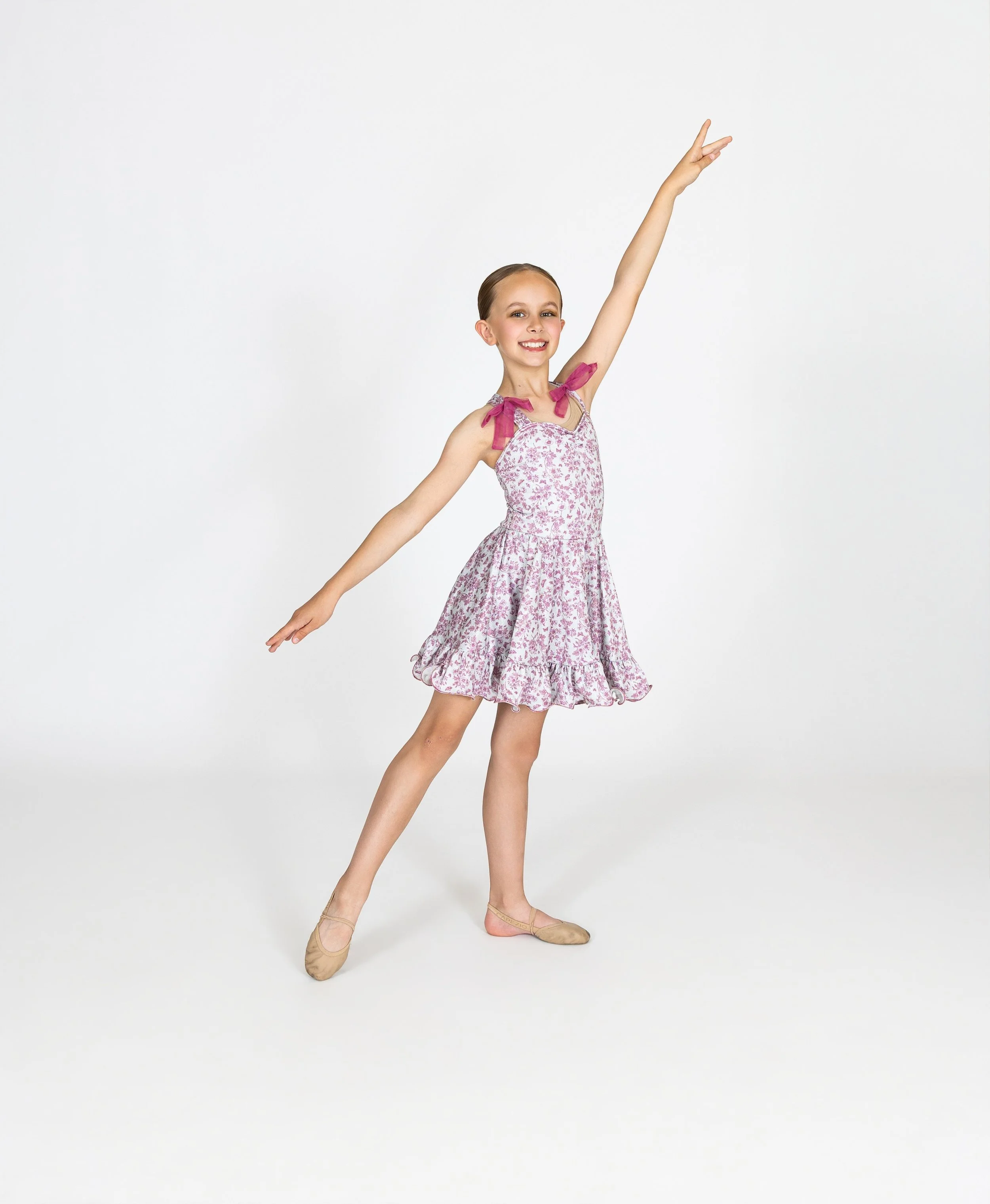 Dancer in a pink floral dance dress and ballet shoes, performing a ballet pose with her right arm raised and her left arm extended sideways, standing on a white background.
