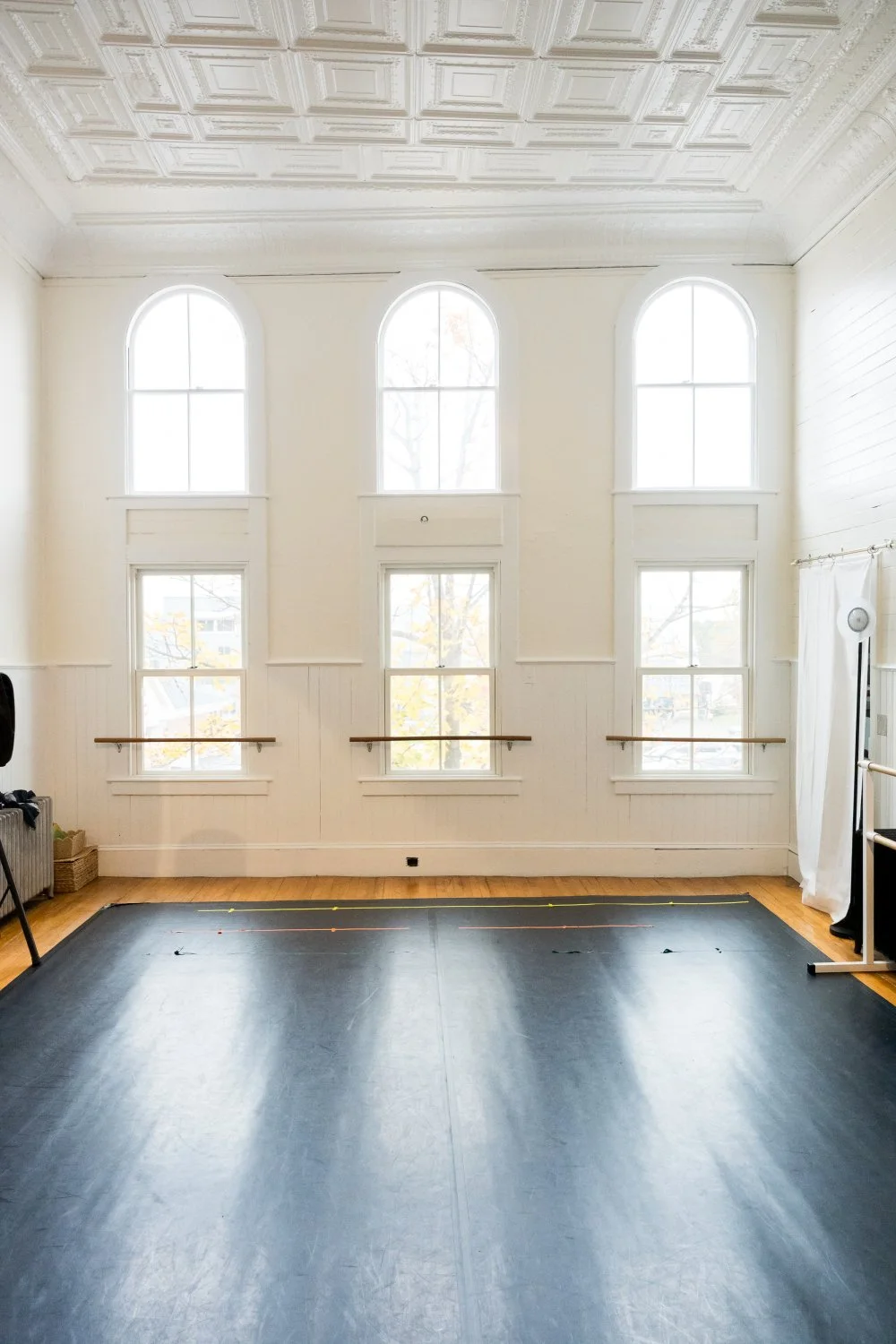 Empty dance studio with large arched windows, white walls, wooden flooring, and a black dance mat in the center.