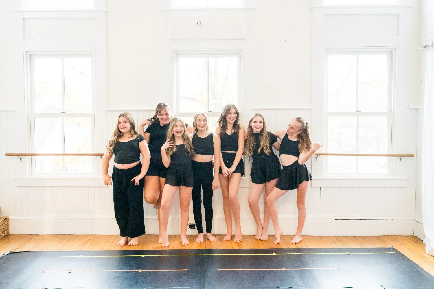 Dancers standing in a brightly lit dance studio, smiling and posing together.